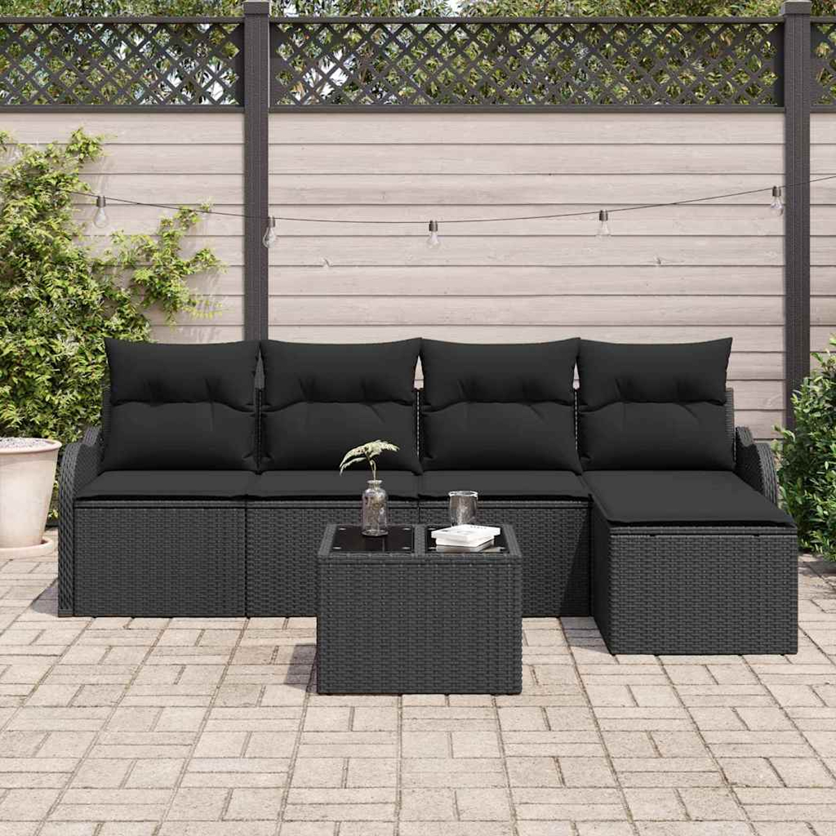 Ein schwarzes Rattan-Sofa mit schwarzen Kissen und ein passender Tisch stehen auf einer Ziegelterrasse. Dahinter ist ein Holzzaun.