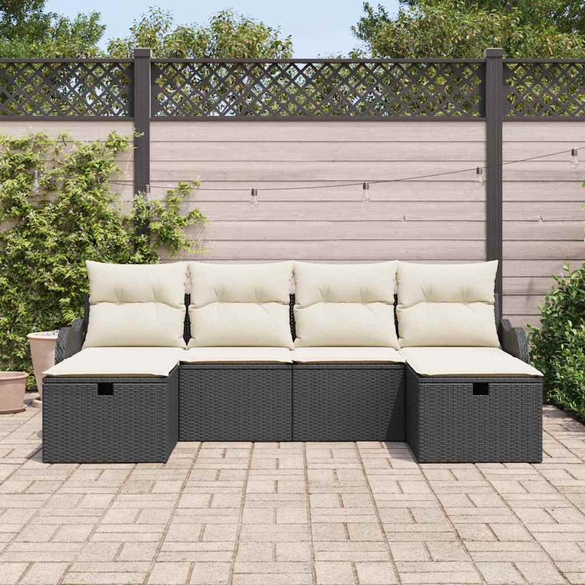 Dunkelgraues Outdoor-Ecksofa mit cremefarbenen Kissen auf einer Terrasse. Im Hintergrund ein Holzzaun mit Grün und Lichterketten.