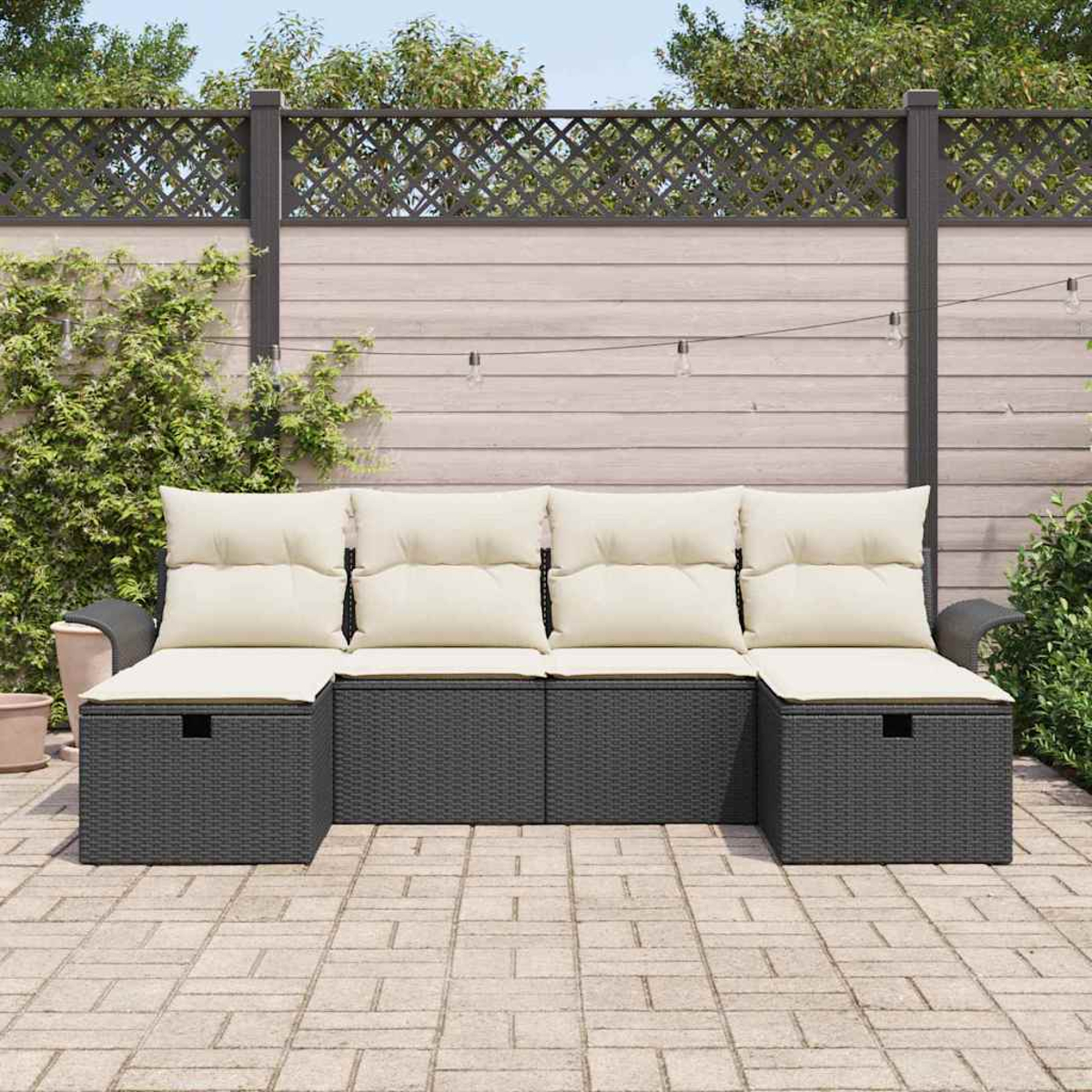 Outdoor-Ecksofa mit cremefarbenen Kissen und dunkelgrauem Korbgeflecht. Im Hintergrund ein Holzzaun mit Pflanzen, Lichterketten und eine Ziegelterrasse.