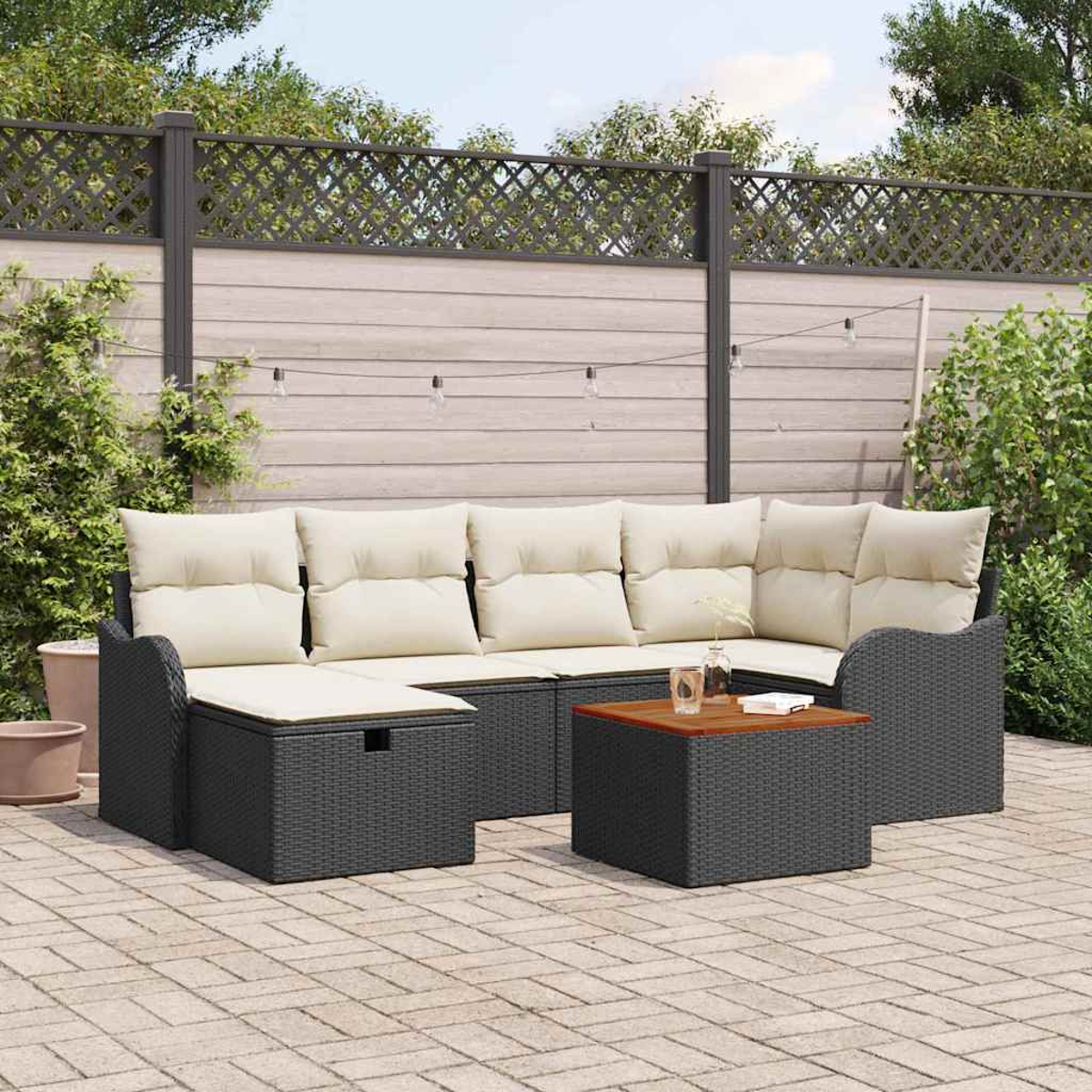 Outdoor-Ecksofa mit cremefarbenen Kissen und einem kleinen Tisch. Im Hintergrund ein Zaun und Grün, auf einer gepflasterten Terrasse.