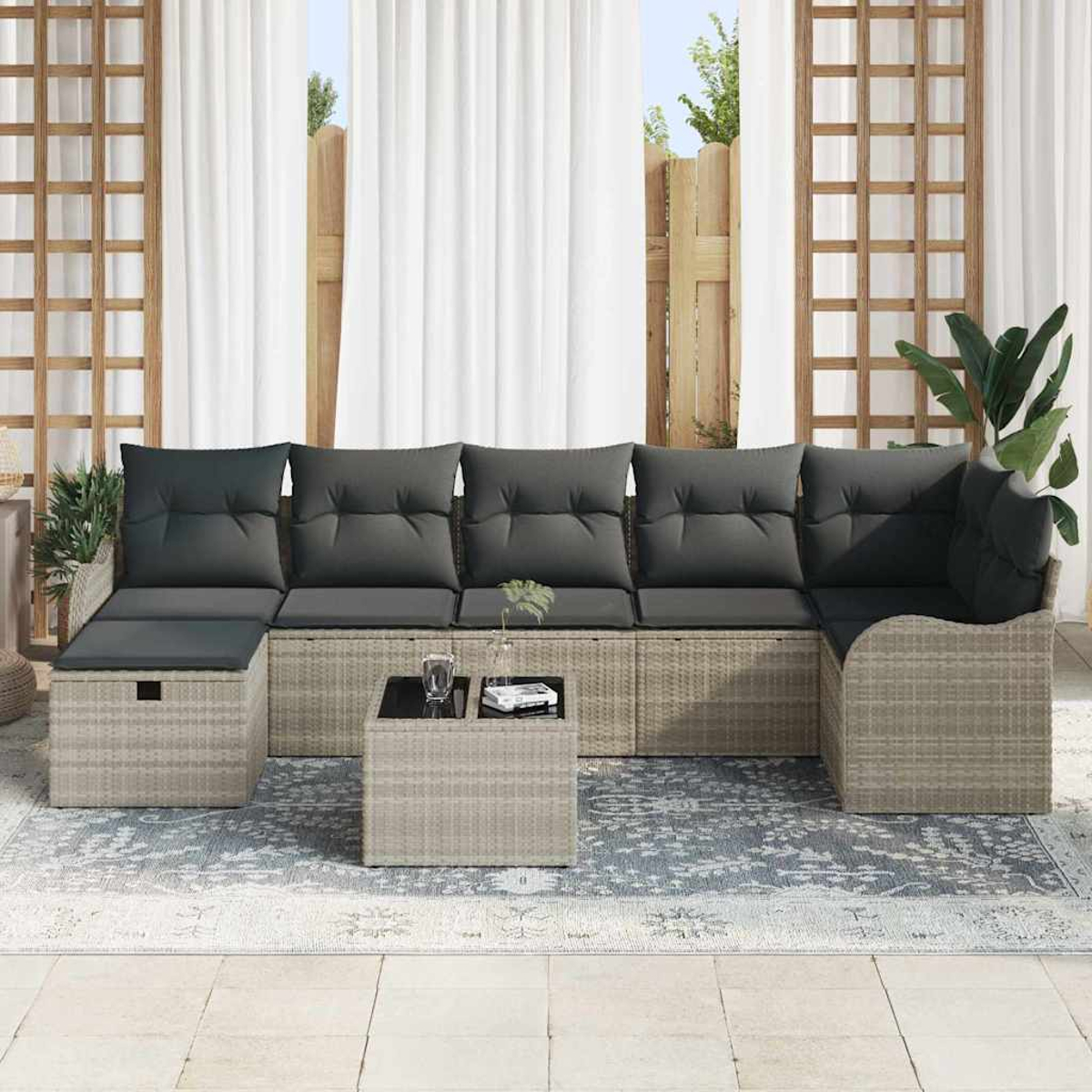 Ein graues Ecksofa im Freien mit schwarzen Kissen, einem kleinen Tisch und einem Teppich. Weiße Vorhänge und Holzgitter im Hintergrund.