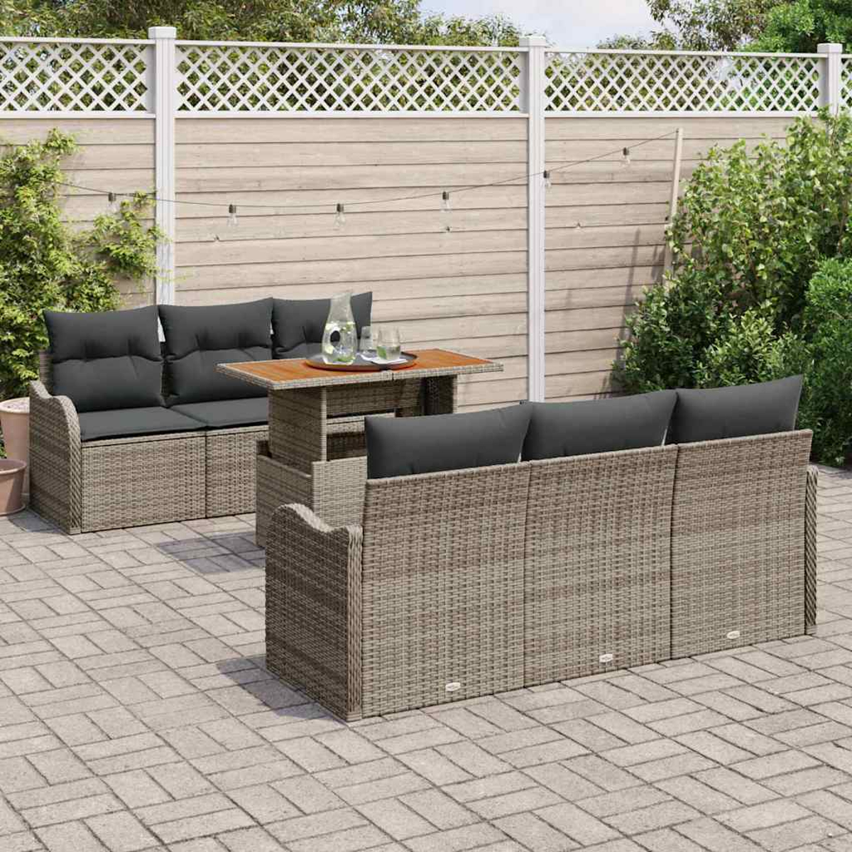 Juego de patio exterior: sofás de mimbre con cojines grises, mesa de madera y una valla al fondo. La escena tiene lugar en un patio pavimentado.