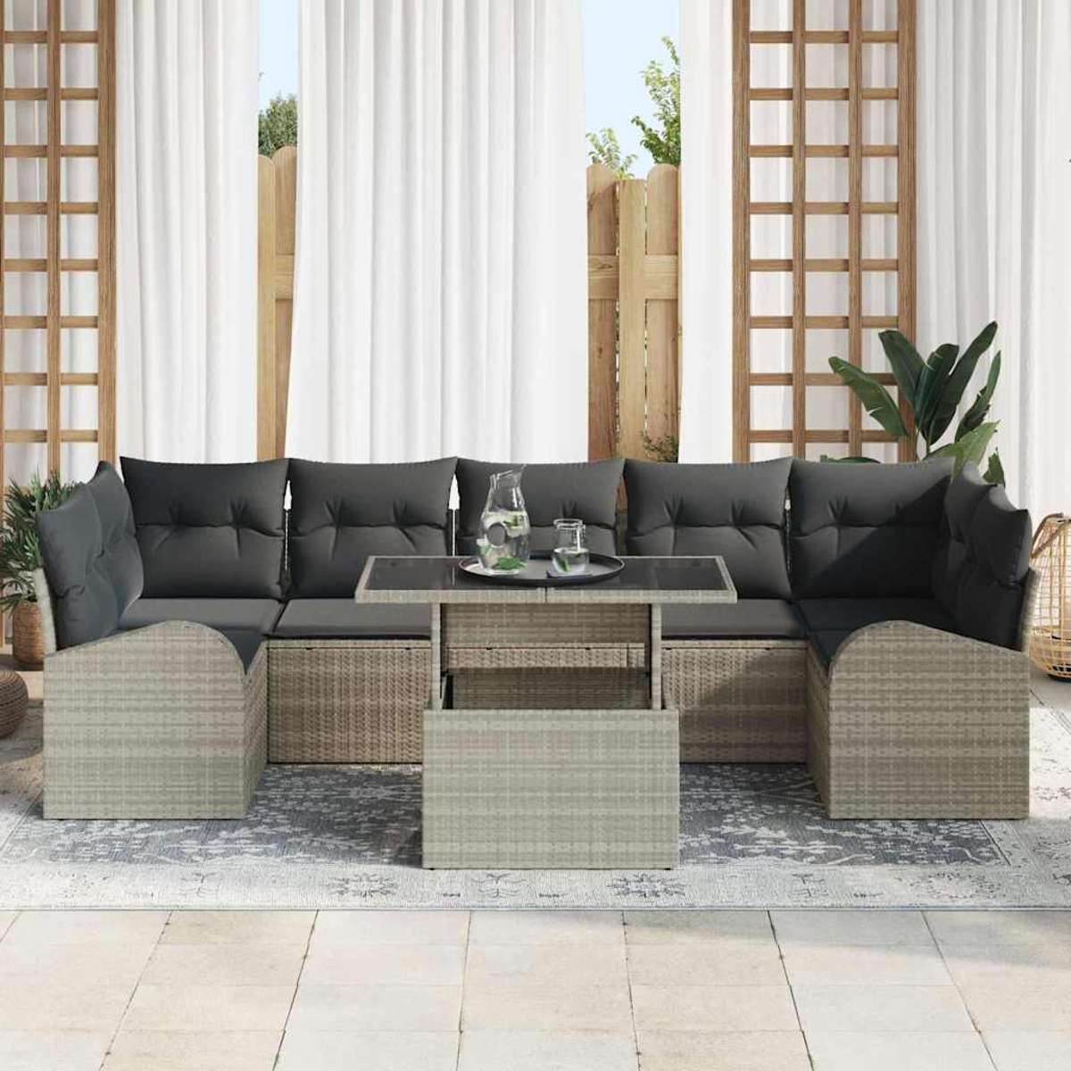 Outdoor-Rattansofa-Set mit dunkelgrauen Kissen, Glastisch und weißen Vorhängen im Hintergrund. Pflanze und Spalier sichtbar.