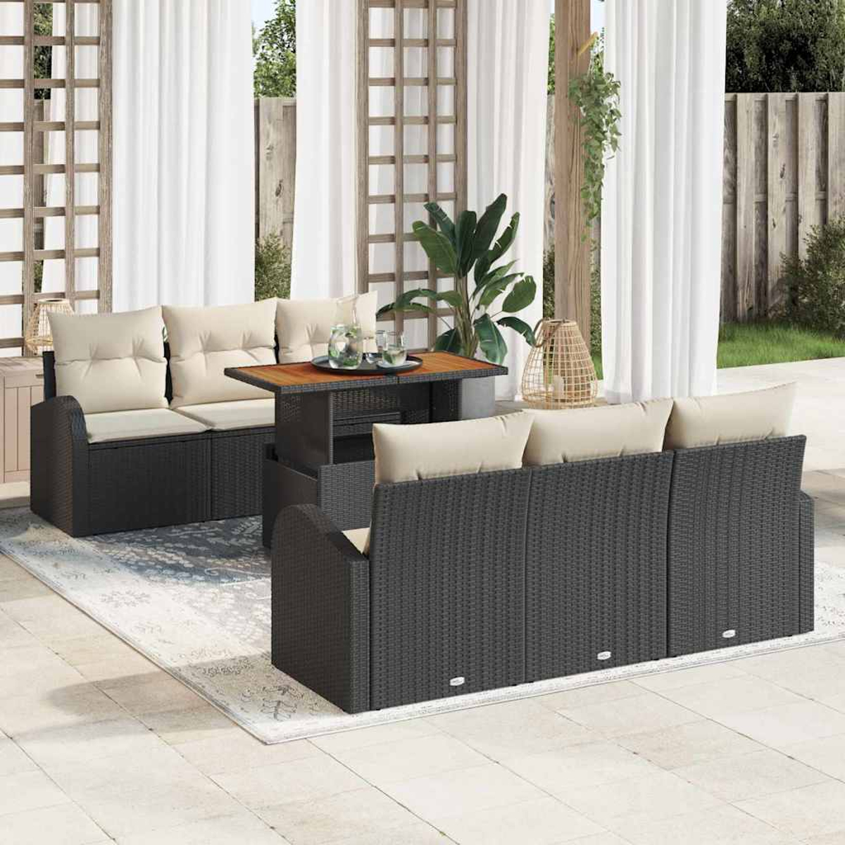 Juego de patio exterior con muebles de mimbre oscuro, cojines beige, mesa de madera, cortinas blancas y una alfombra. El fondo muestra una valla de madera.