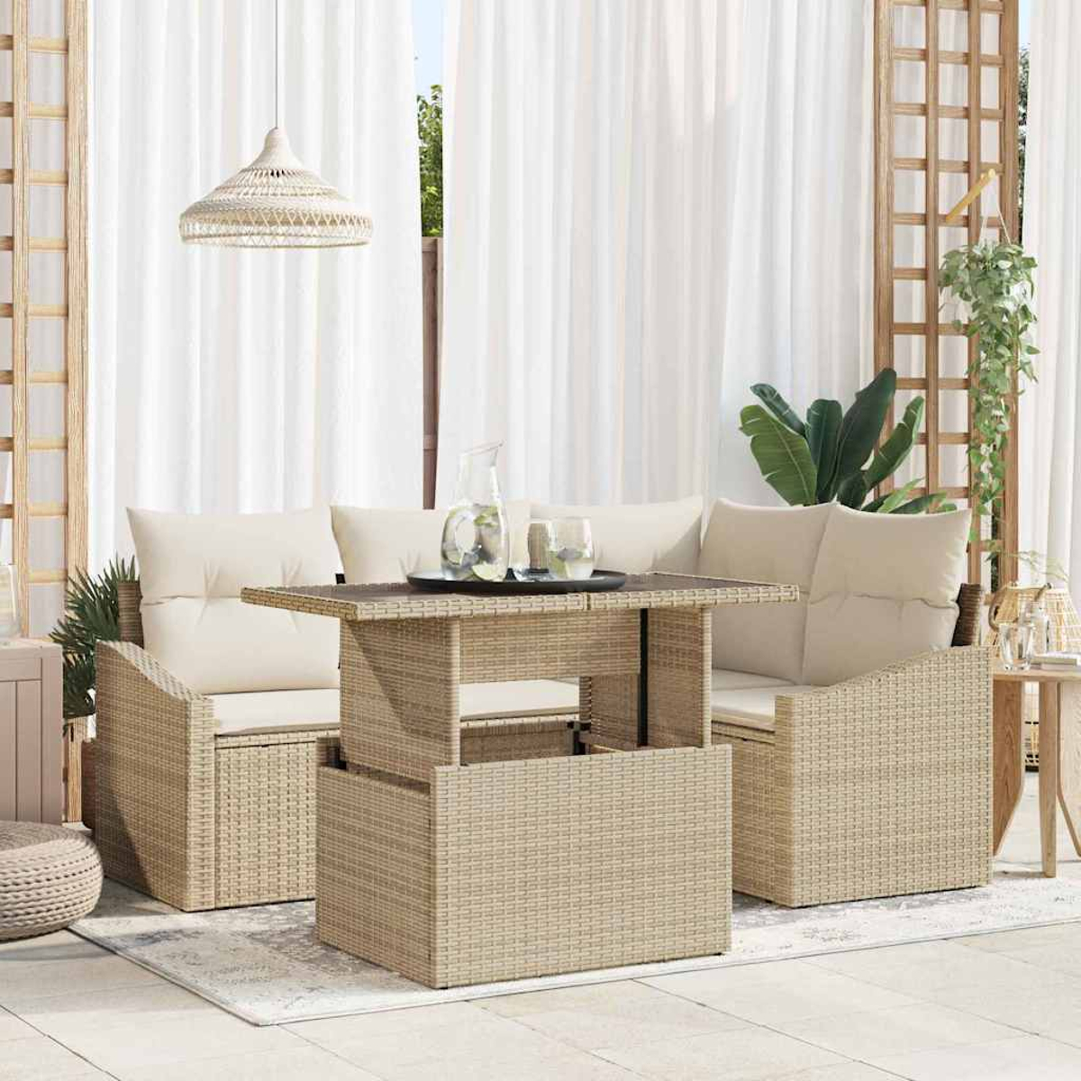 Geflochtenes Terrassenmöbel-Set mit Tisch, Kissen und Pendelleuchte. Weiße Vorhänge und Grünzeug im Hintergrund.