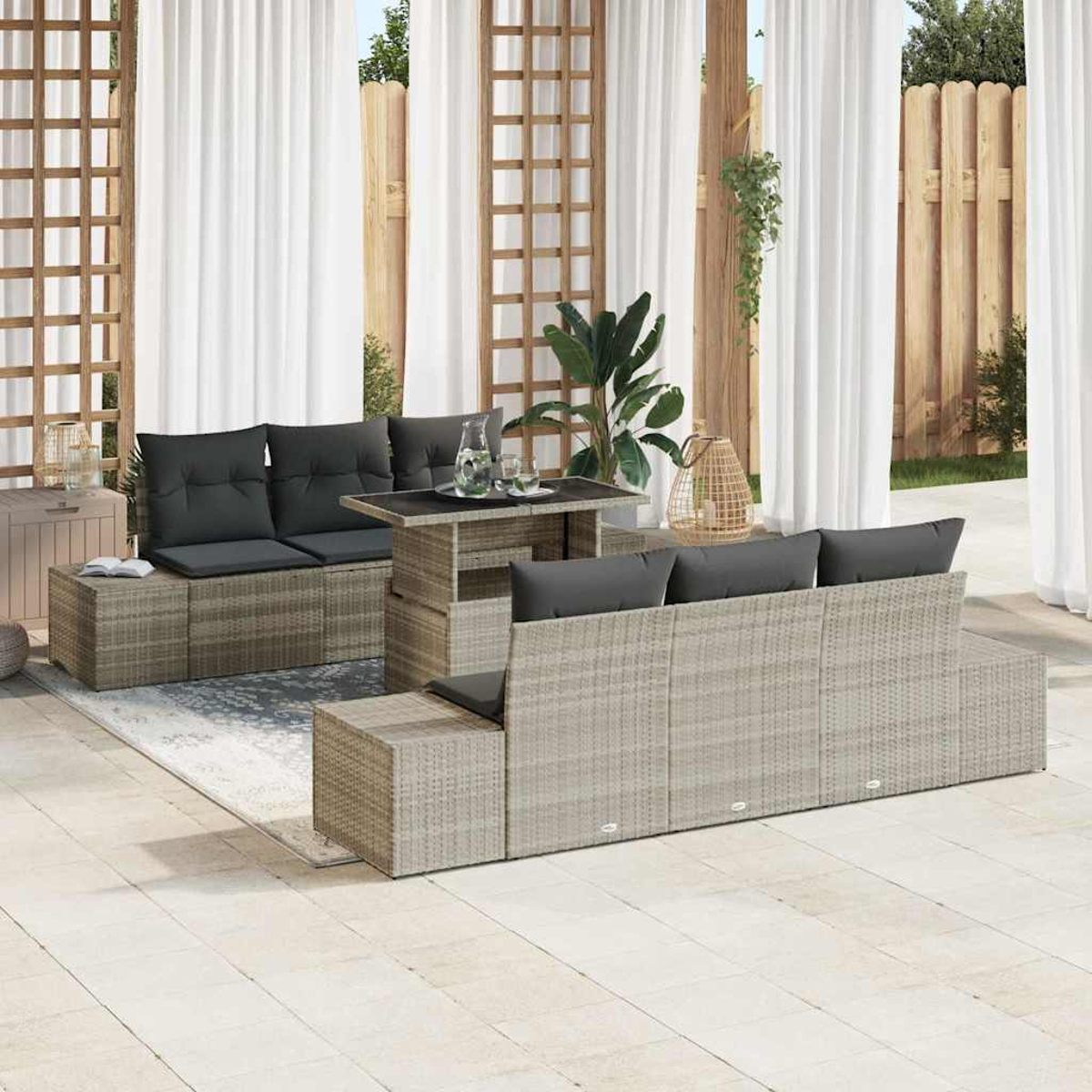 Mobiliario de mimbre para exteriores con cojines grises, mesa y plantas. Cortinas blancas y enrejado de madera al fondo.
