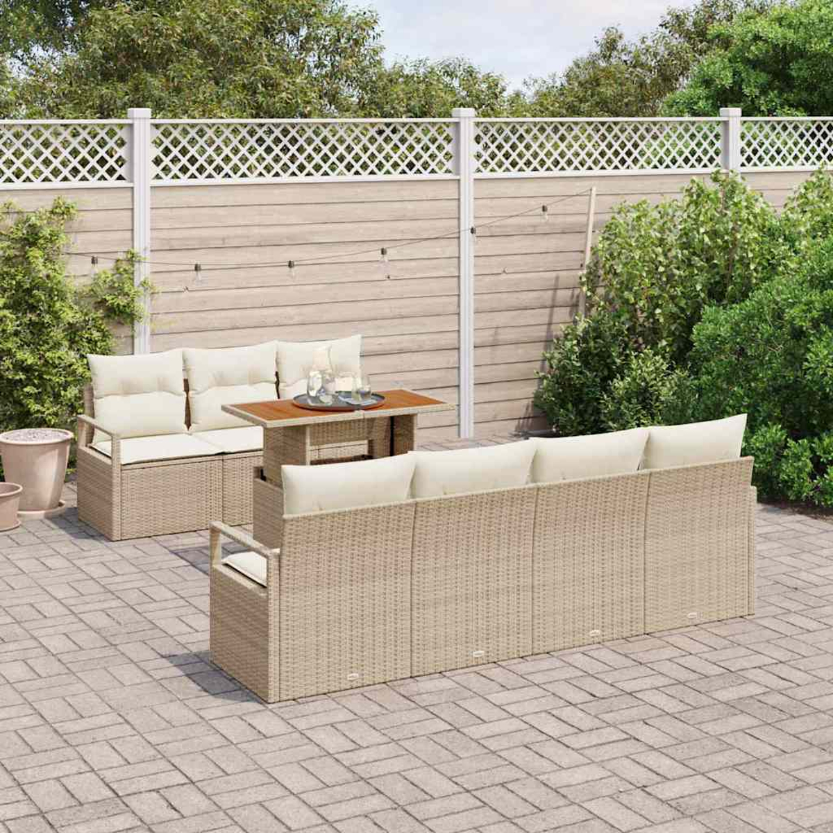 Garten-Set mit beigen Korbmöbeln, cremefarbenen Kissen und einem Holztisch. Ein weißer Zaun und grüne Büsche im Hintergrund.