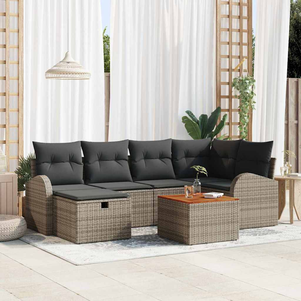 Zona de estar al aire libre con cojines grises, muebles de mimbre y una mesa de madera. Cortinas blancas y plantas verdes al fondo.