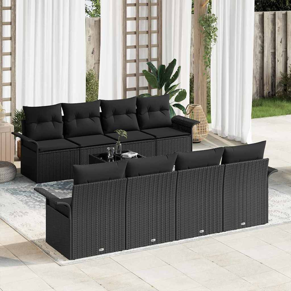 Escena al aire libre con muebles de mimbre negro. Incluye un sofá, un sofá de dos plazas y una mesa de centro. Cortinas blancas y vegetación al fondo.