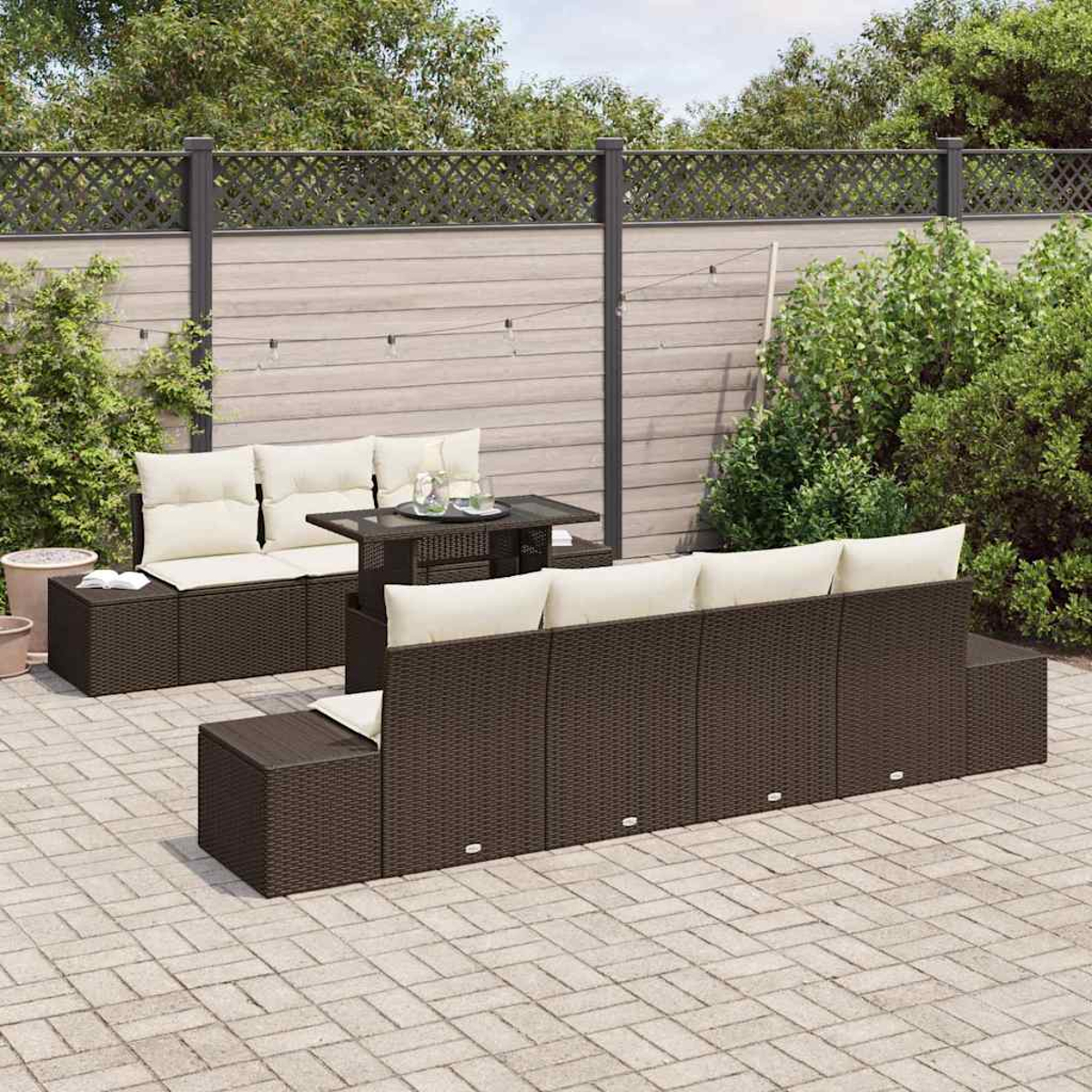 Garten-Sitzgruppe mit braunen Korbmöbeln, weißen Kissen und einem Tisch mit Glasplatte auf einer Ziegelterrasse, im Hintergrund ein Zaun.