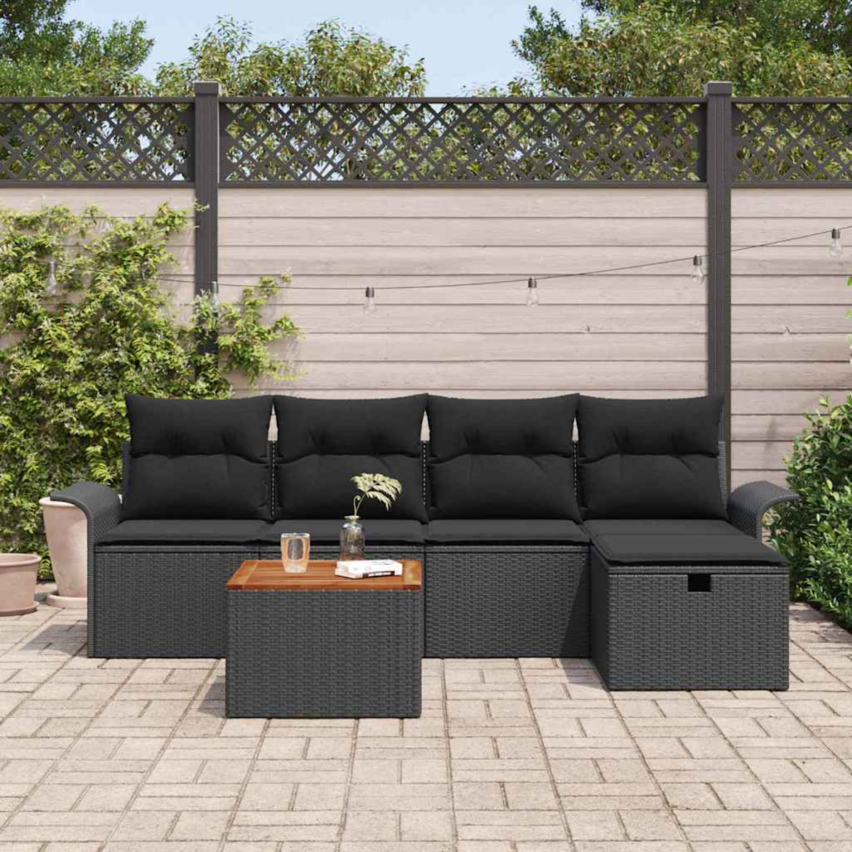 Szene im Freien mit einem grauen Rattan-Ecksofa, einem Holztisch und einer Ziegelterrasse. Ein dekorativer Zaun und grüne Pflanzen sind im Hintergrund.