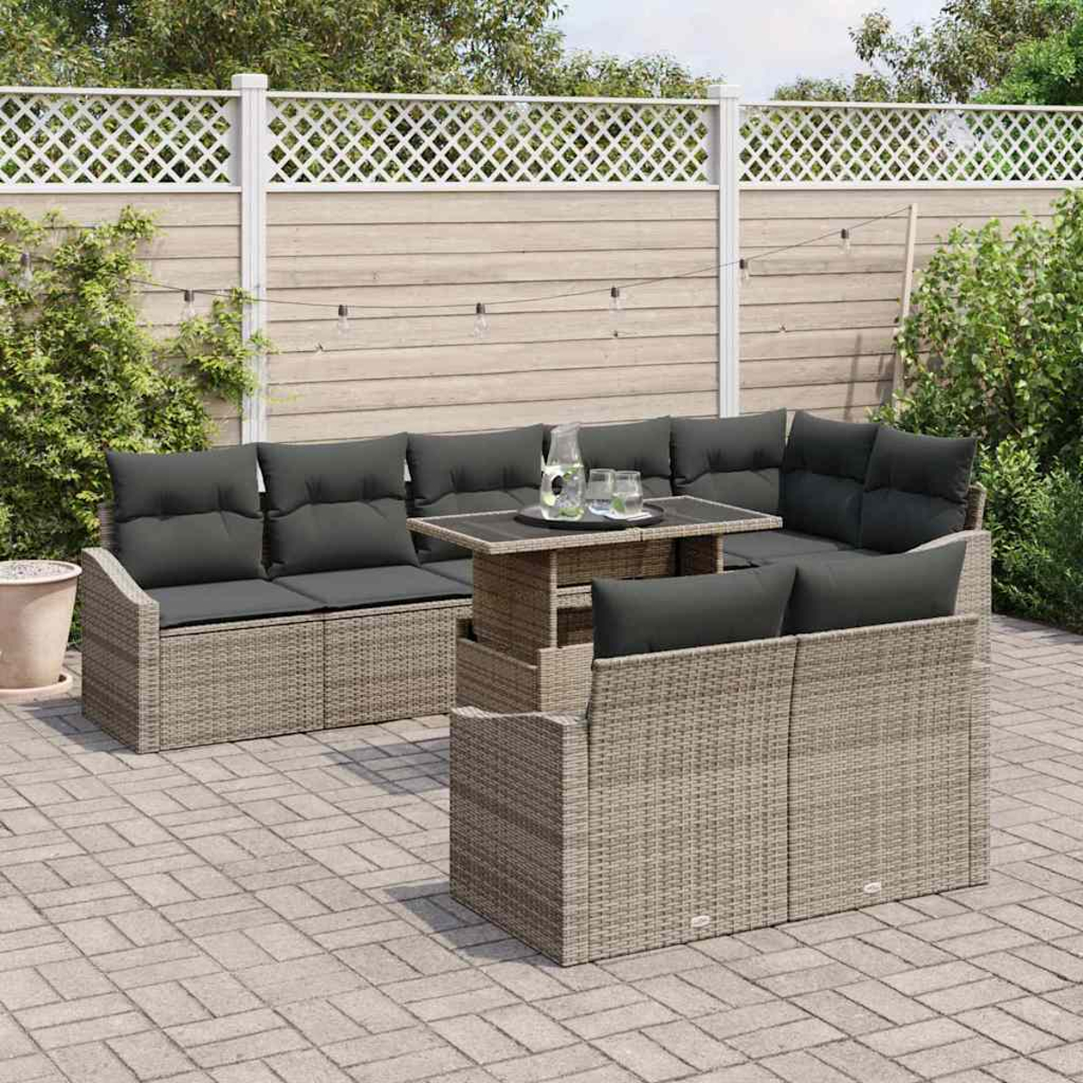 Gartenmöbel-Set aus Korbgeflecht mit grauen Kissen und einem Tisch auf einer Steinterrasse, mit weißem Zaun im Hintergrund.
