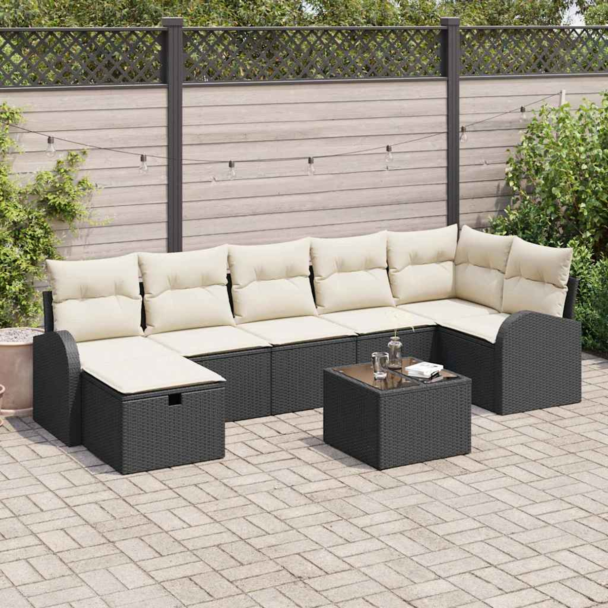 Sofá seccional exterior con cojines crema, mesa y valla decorativa. El patio está embaldosado. Hay plantas y una guirnalda de luces.