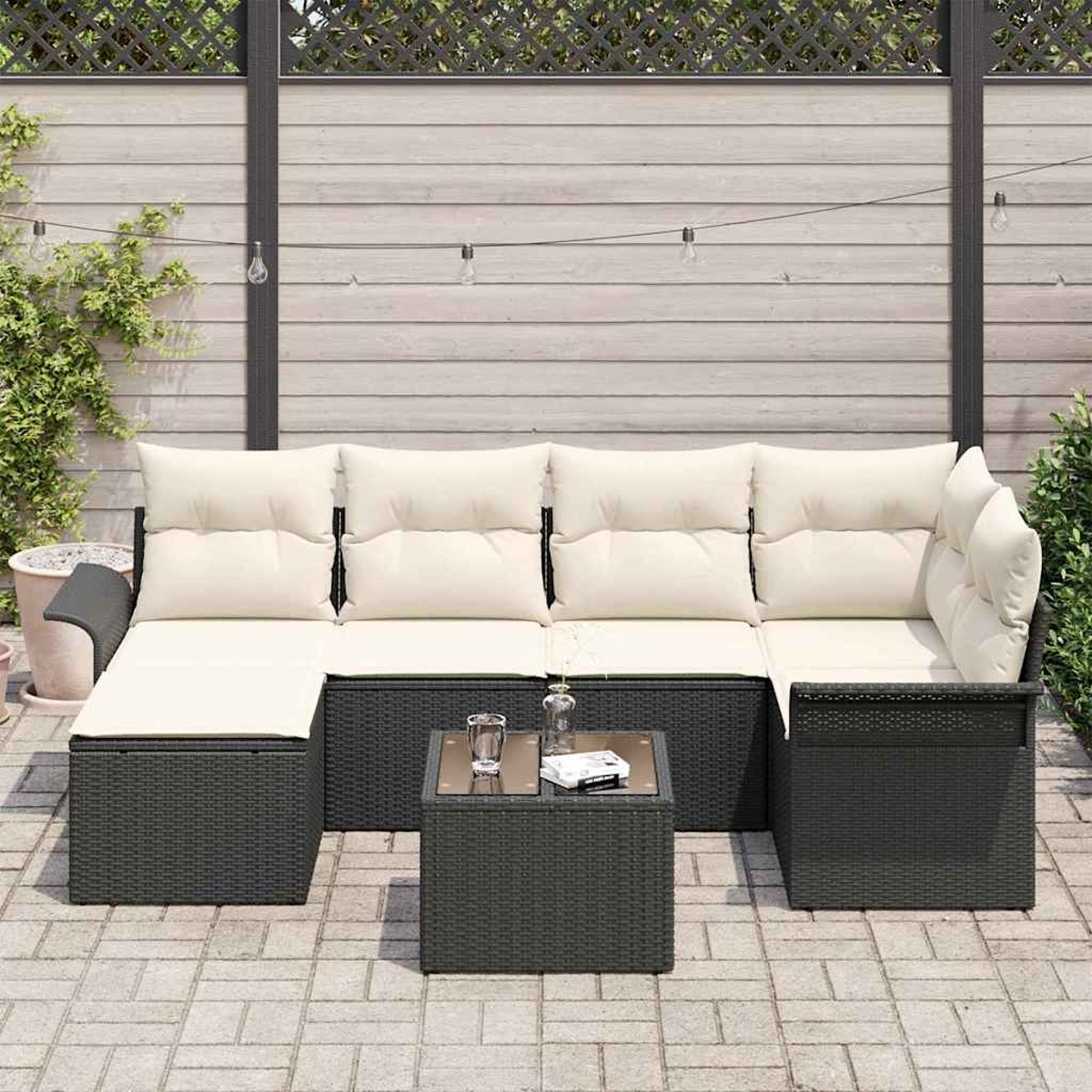 Dunkelgraues Outdoor-Ecksofa aus Korbgeflecht mit cremefarbenen Kissen und einem kleinen quadratischen Tisch mit Glasplatte. Im Hintergrund ein Holzzaun.
