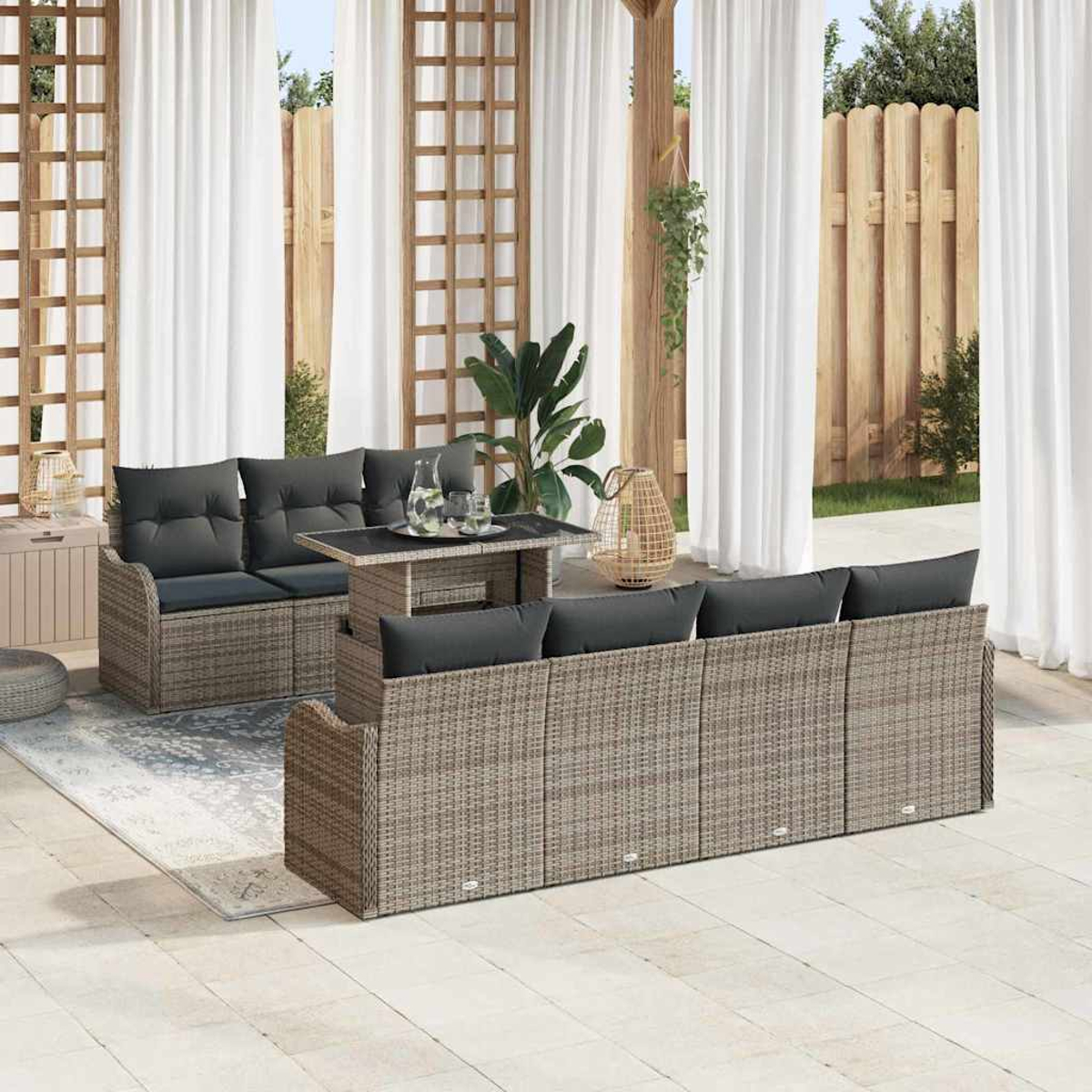 Terrassenset mit grauen Kissen, Tisch mit Gläsern und weißen Vorhängen im Hintergrund. Holzzaun und grüne Pflanzen.