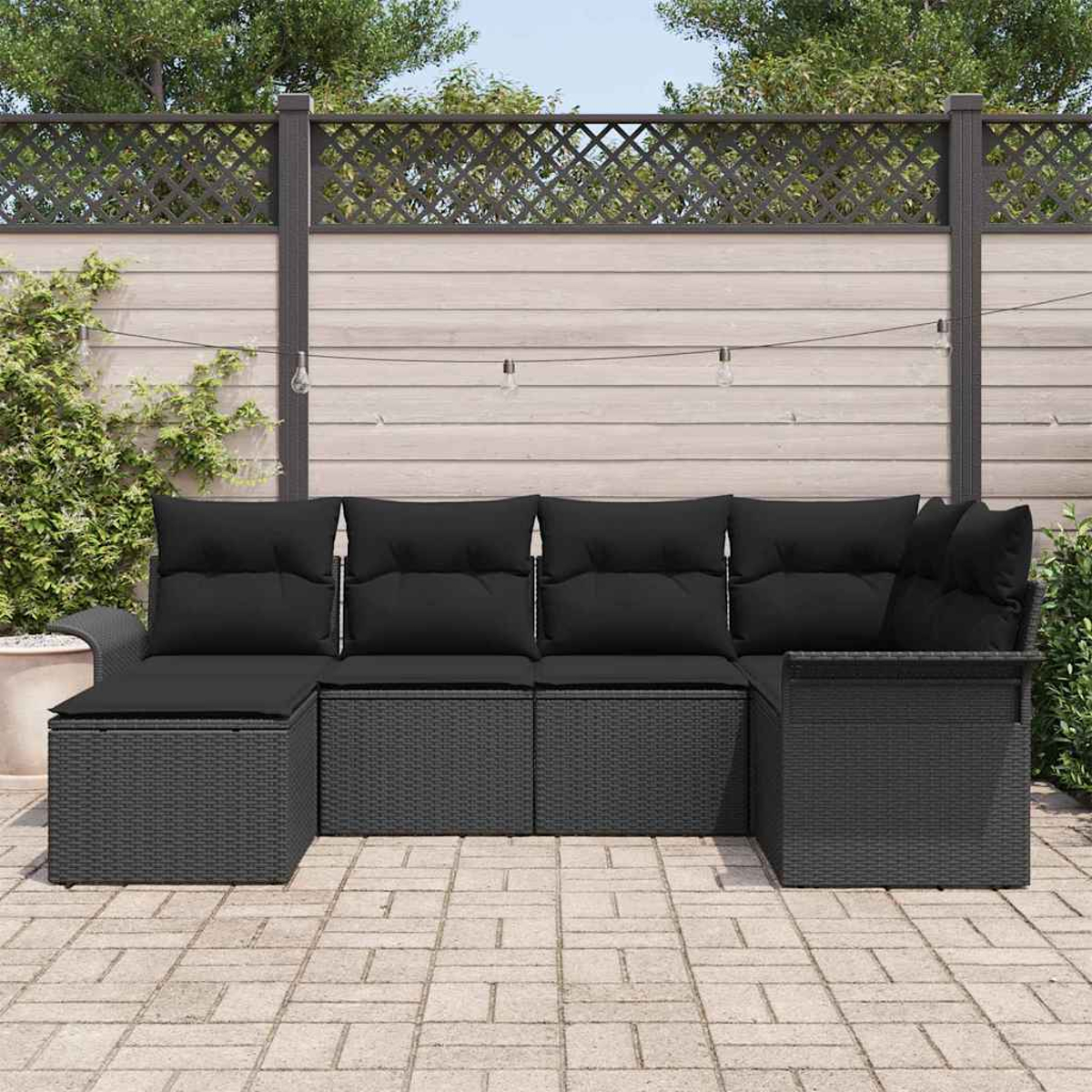 Ein schwarzes Outdoor-Ecksofa aus Korbgeflecht mit schwarzen Kissen steht auf einer Steinpflasterterrasse. Ein Holzzaun befindet sich im Hintergrund.