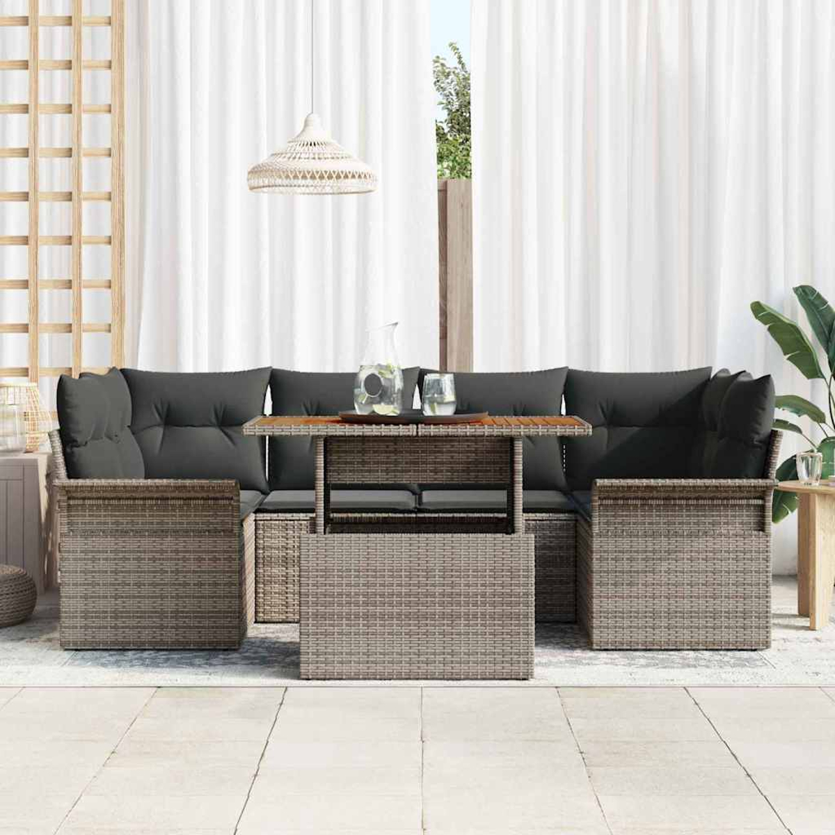 Gartenmöbel-Set aus Korbgeflecht mit grauen Kissen, einem Tisch mit Glaskrug und zwei Gläsern sowie weißen Vorhängen im Hintergrund.