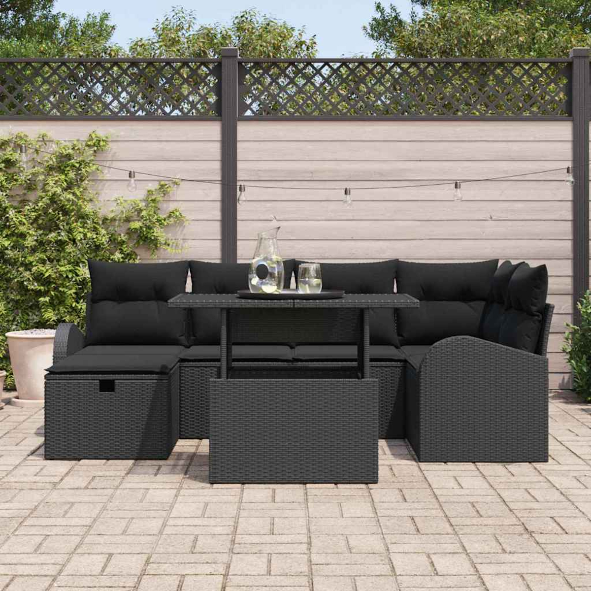 Schwarzes Outdoor-Wickermöbelset mit Kissen, Tisch und Wasserkrug. Ein Zaun und Grün im Hintergrund.