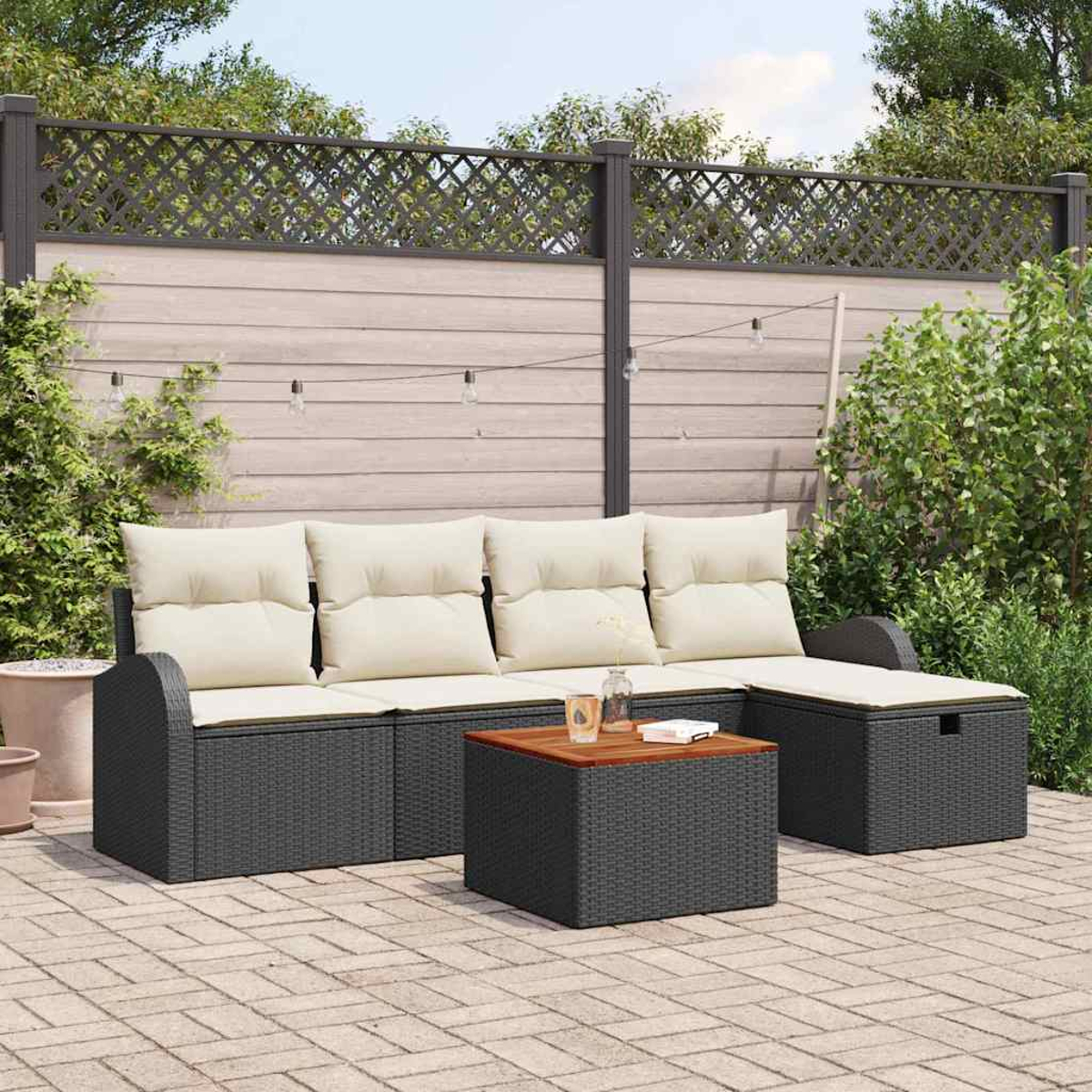 Juego de patio al aire libre con sofá de mimbre gris oscuro, cojines beige y mesa de madera. Una valla y vegetación al fondo.