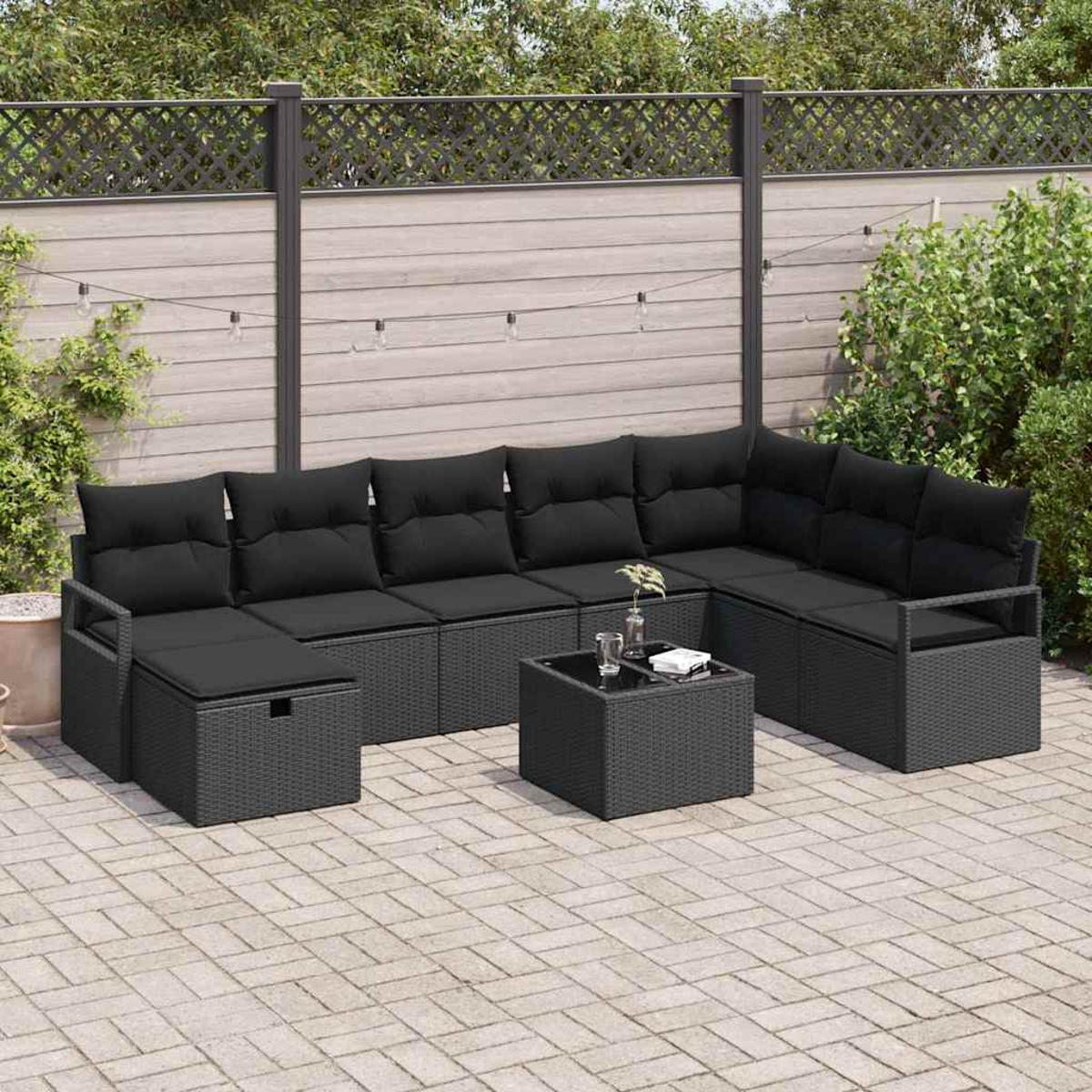 Outdoor-Ecksofa mit schwarzen Kissen auf einer Terrasse. Ein kleiner Tisch mit Gläsern und einer Pflanze steht davor. Hintergrund Zaun und Pflanzen.