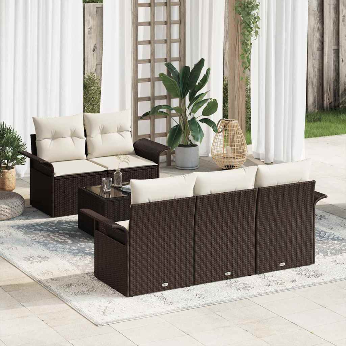 Garten-Set mit braunen Korbmöbeln und cremefarbenen Kissen. Ein Tisch mit Glasplatte, Teppich, Pflanzen und weiße Vorhänge.