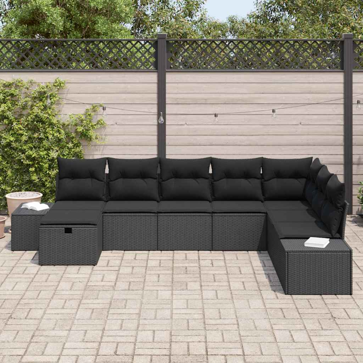 Outdoor-Ecksofa mit schwarzen Kissen. Grauer Korbrahmen auf einer Ziegelterrasse, im Hintergrund ein Holzzaun.
