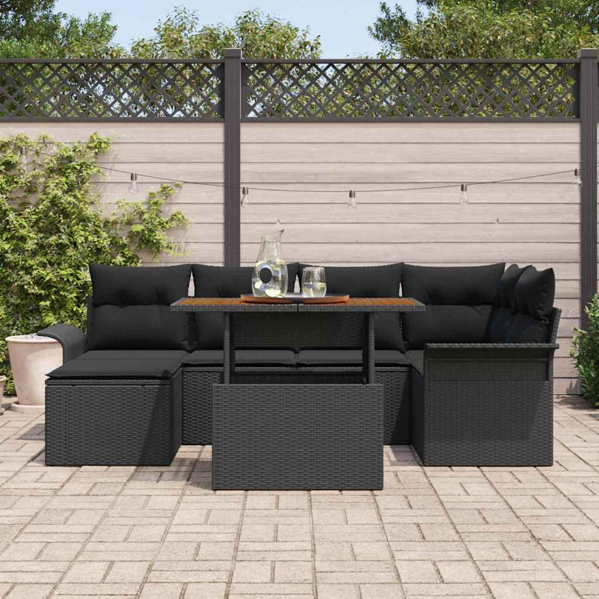 Garten-Sitzgruppe auf einer Terrasse. Schwarze Korbmöbel mit schwarzen Kissen und einem Holztisch. Eine durchsichtige Kanne und Gläser stehen auf dem Tisch.