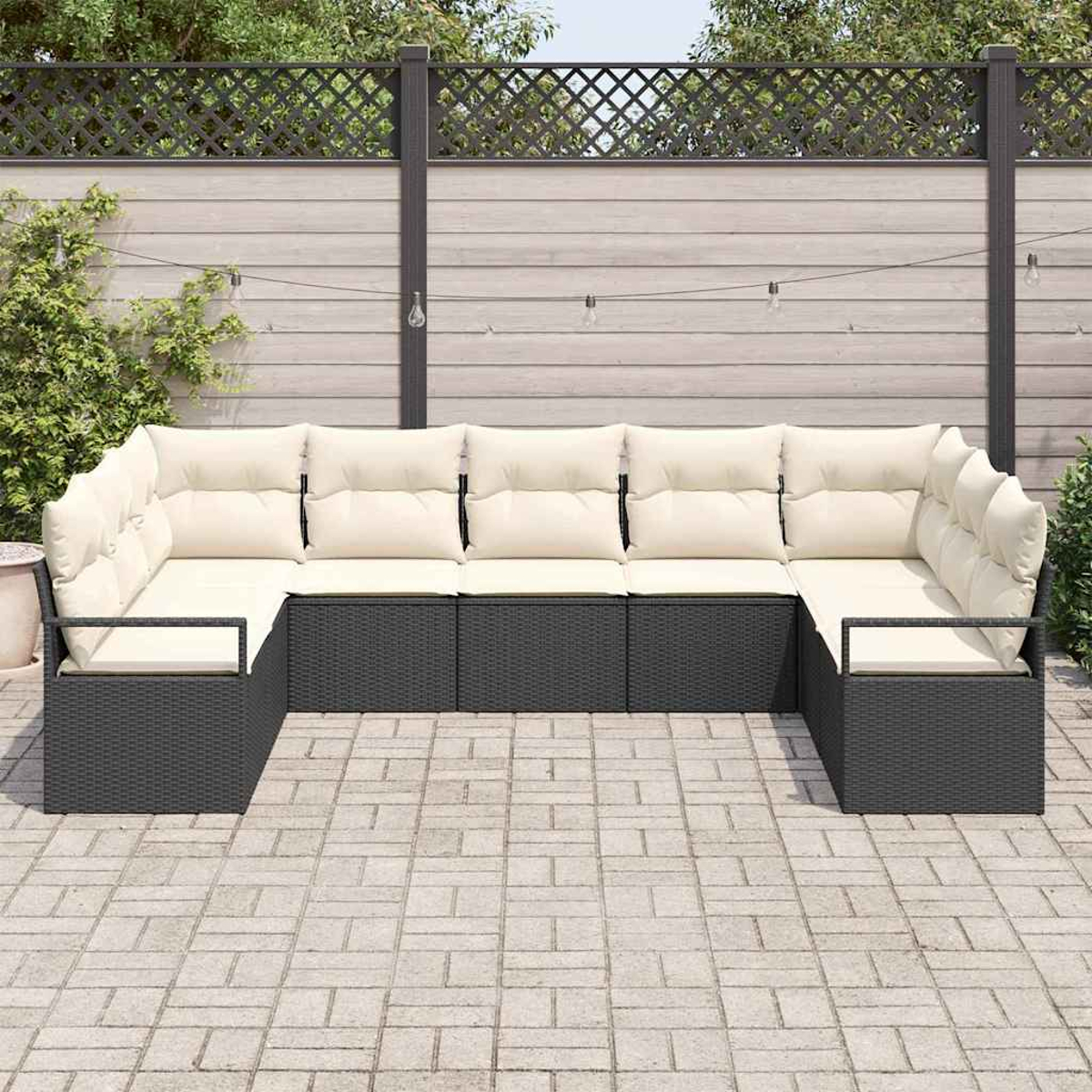 Ein U-förmiges Outdoor-Sofa mit cremefarbenen Kissen steht auf einer Backstein-Terrasse. Im Hintergrund befinden sich ein Zaun mit Lichterketten und Grün.