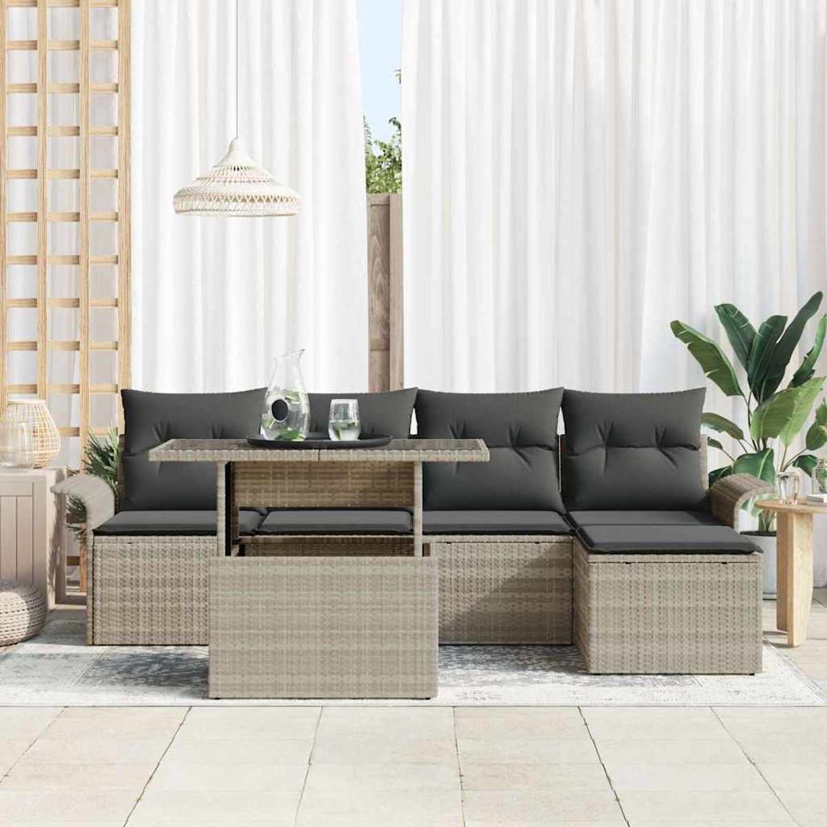 Zona de estar al aire libre con cojines grises, una mesa de cristal y una lámpara tejida. Cortinas y plantas al fondo.