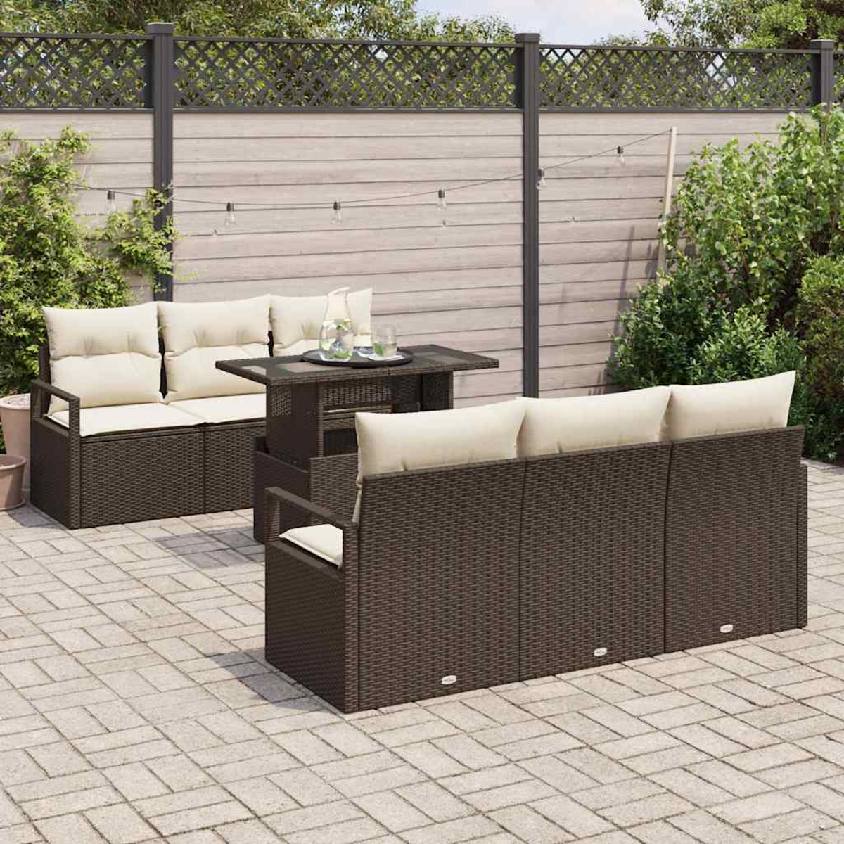 Terrassenset mit braunen Korbmöbeln, cremefarbenen Kissen und einem dunklen Tisch. Im Hintergrund ein Zaun und Grün.