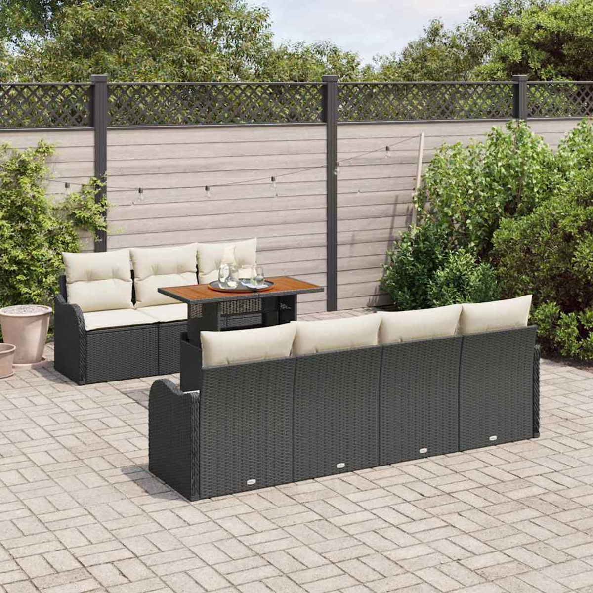 Garten-Set mit zwei Sofas, einem Tisch und beigefarbenen Kissen. Auf Steinpflaster mit Holzzaun und Grün im Hintergrund.
