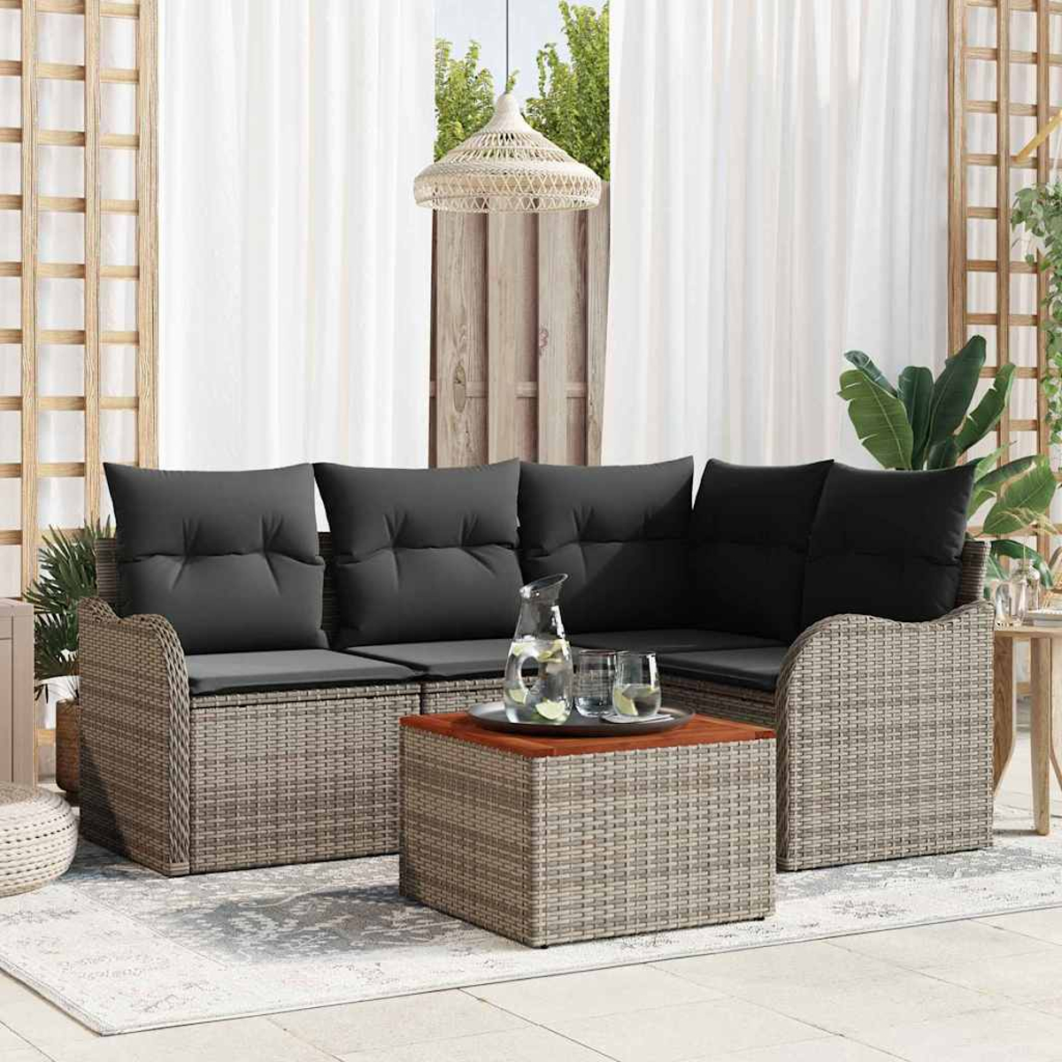 Garten-Sitzecke mit grauen Kissen, Couchtisch und Wasserkrug. Weiße Vorhänge und ein Holzgitter im Hintergrund.