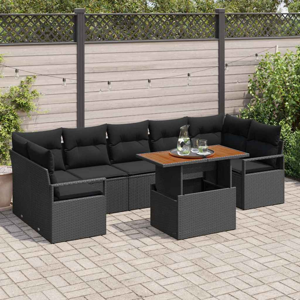 Garten-Terrassenset mit schwarzen Kissen. Ein Holztisch mit Tablett, Gläsern und einem Krug. Graue Korbmöbel auf einer Ziegelterrasse.