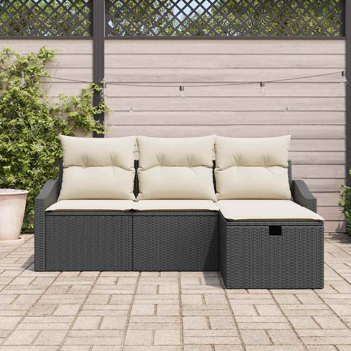 Ein dunkelgraues Outdoor-Sofa aus Korbgeflecht mit cremefarbenen Kissen steht auf einer Steinterrasse. Im Hintergrund befindet sich ein Holzzaun.
