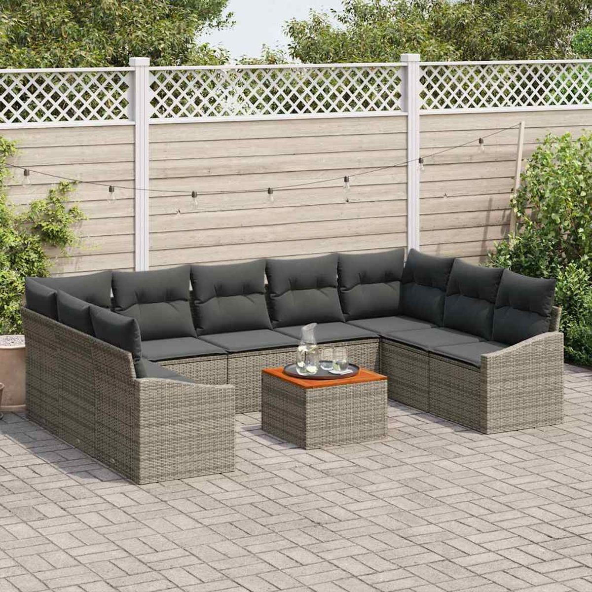 Outdoor-Sitzbereich mit grauen Korbmöbeln, dunklen Kissen und einem kleinen Tisch. Im Hintergrund ein weißer Gitterzaun und Lichterketten.