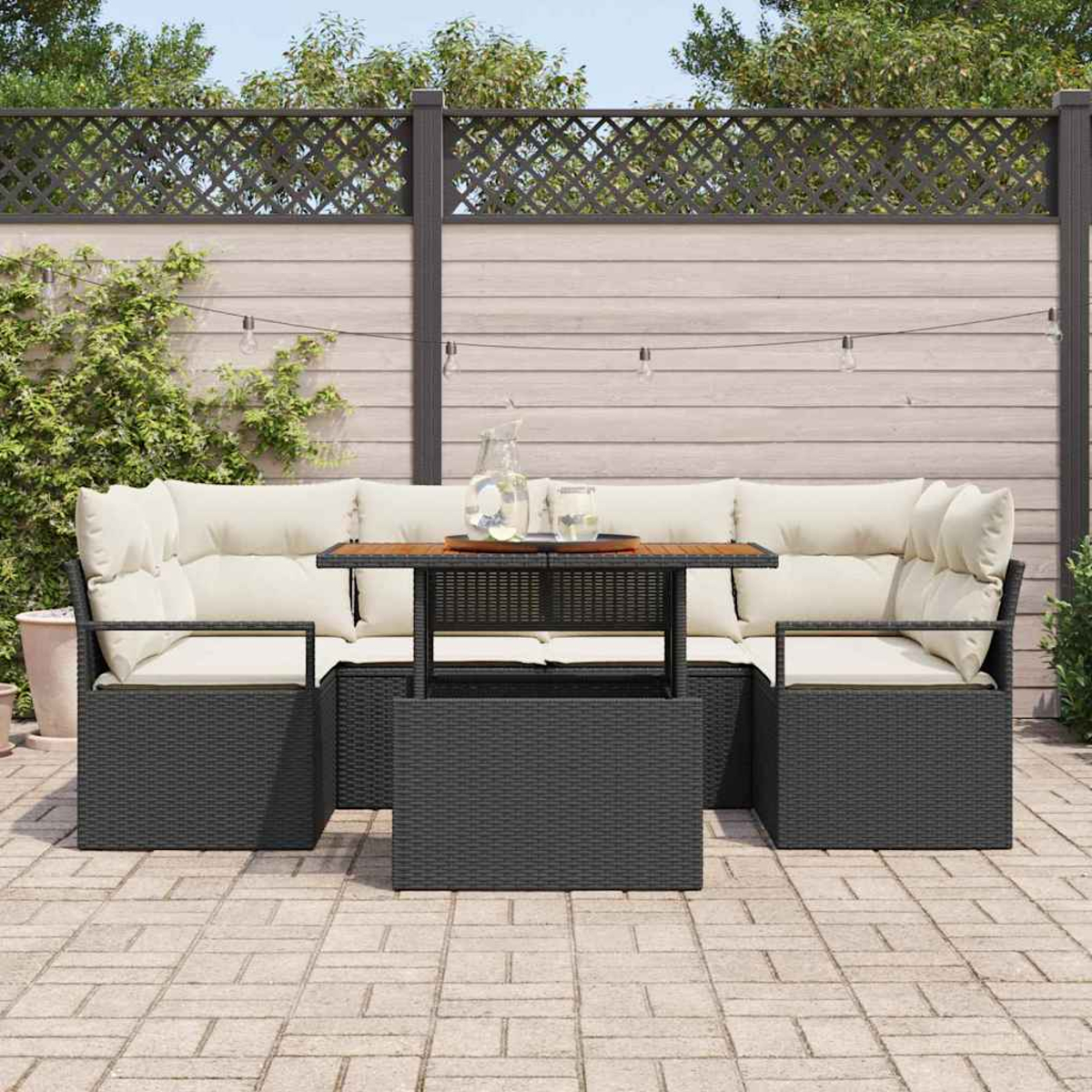 Terrassen-Set mit schwarzer Korbmöbel, beigen Kissen und einem Holztisch. Im Hintergrund befinden sich ein Zaun und Pflanzen.