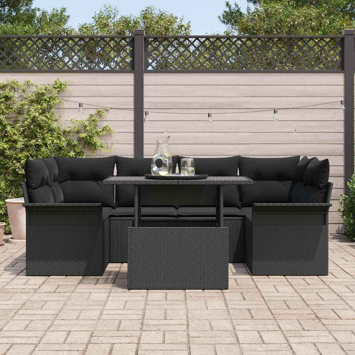 Schwarzes Outdoor-Wicker-Ecksofa mit Kissen, Tisch und Getränken auf einer Terrasse. Ein Holzzaun mit Pflanzen im Hintergrund.