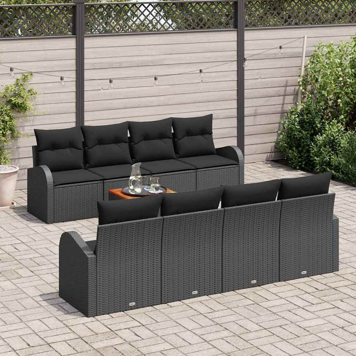 Terrassen-Set mit grauen Korbmöbeln, schwarzen Kissen, einem kleinen Tisch mit Getränken und einem Holzzaun im Hintergrund.