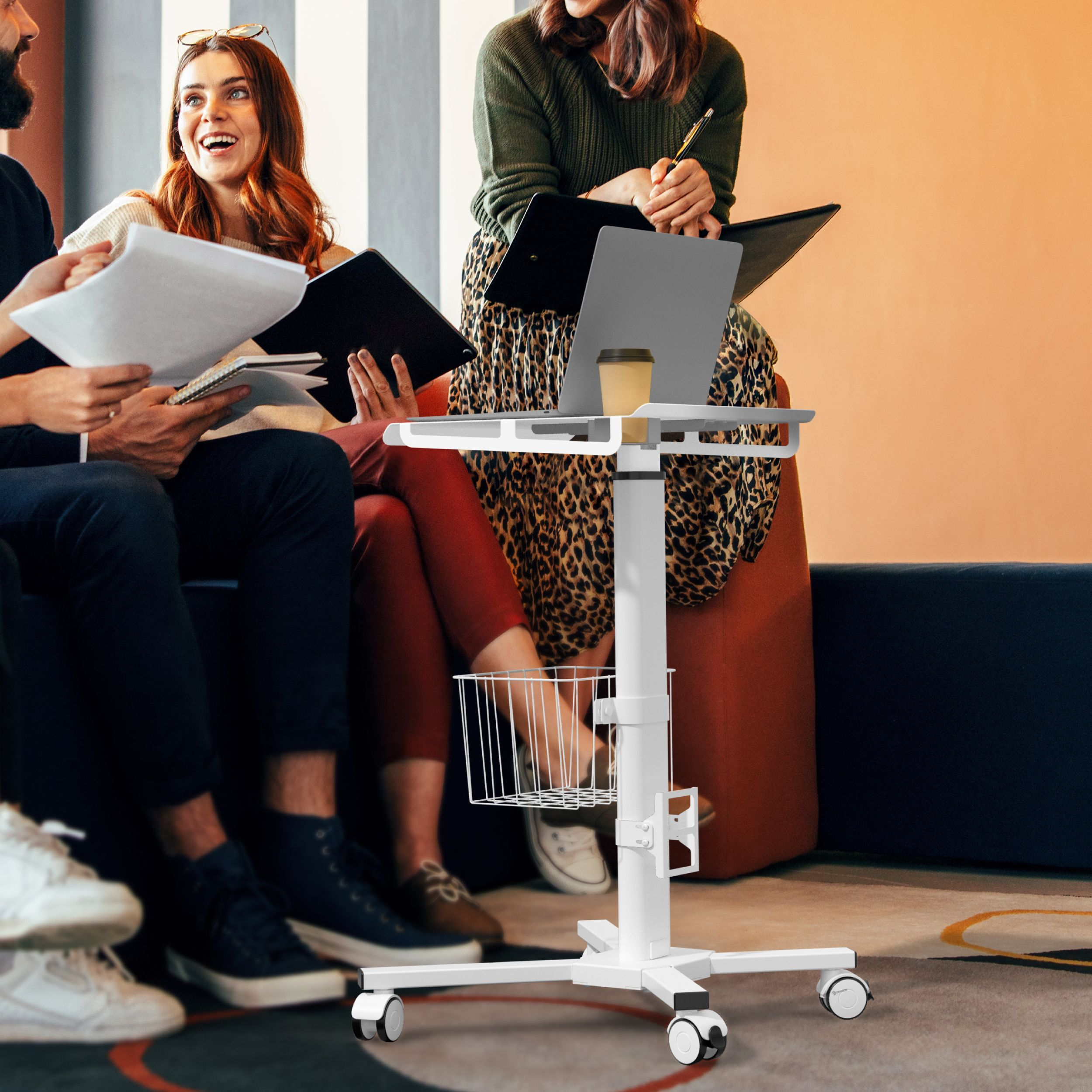 Gruppe von Leuten um einen weißen Rollwagen mit Laptop und Kaffeetasse. Eine Frau lacht, während andere Papiere halten.