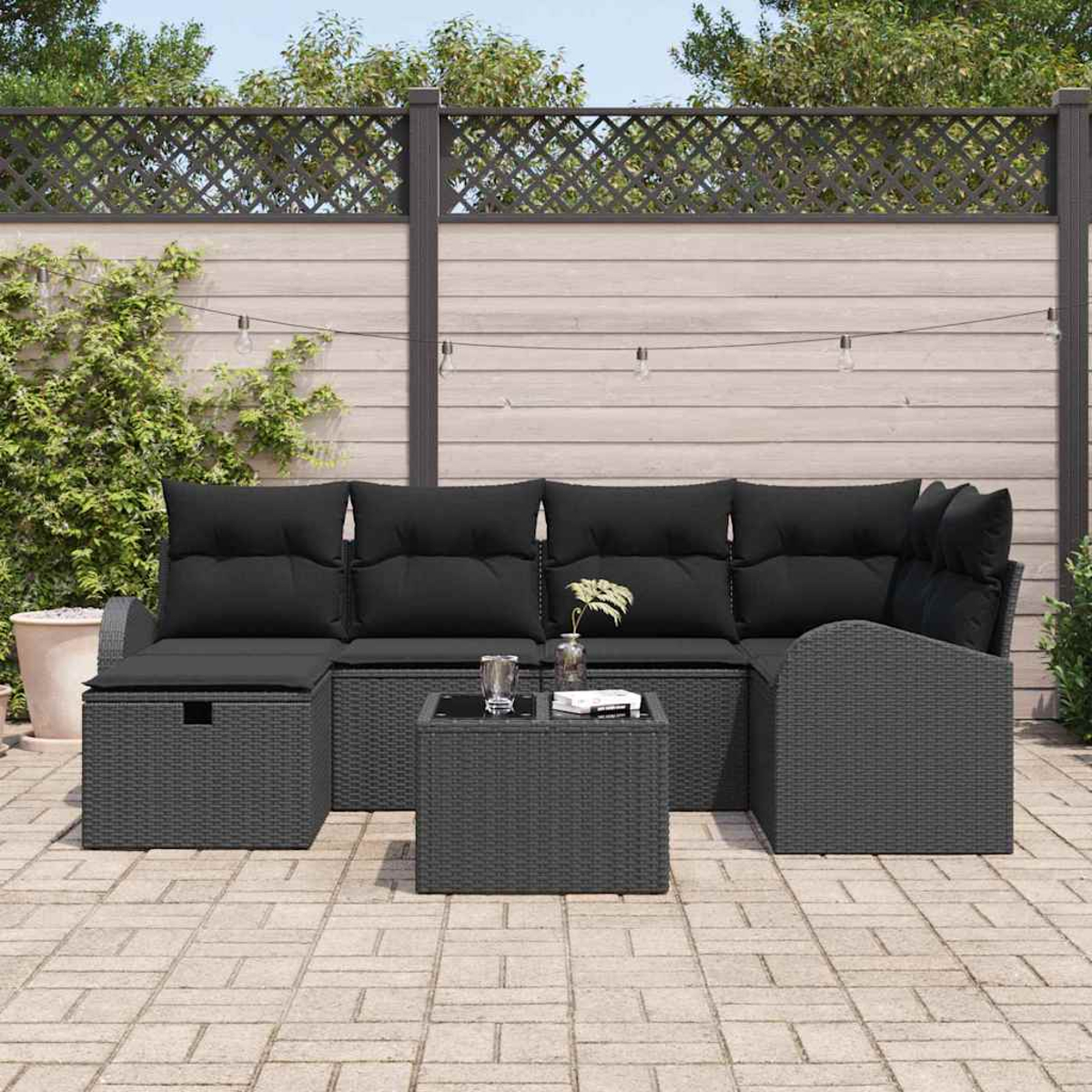 Graues Outdoor-Geflecht-Sofaset mit schwarzen Kissen und einem kleinen Tisch auf einer Backsteinterrasse, im Hintergrund ein Holzzaun.