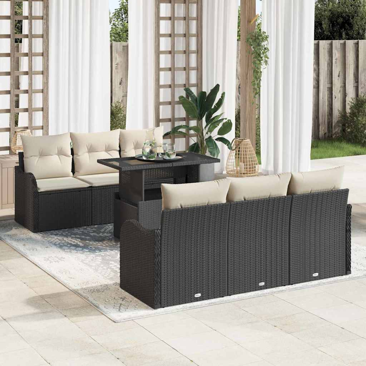 Terrassengarnitur mit dunklem Korbsessel, beigen Kissen und einem Tisch. Transparente Vorhänge und Pflanzen im Hintergrund.