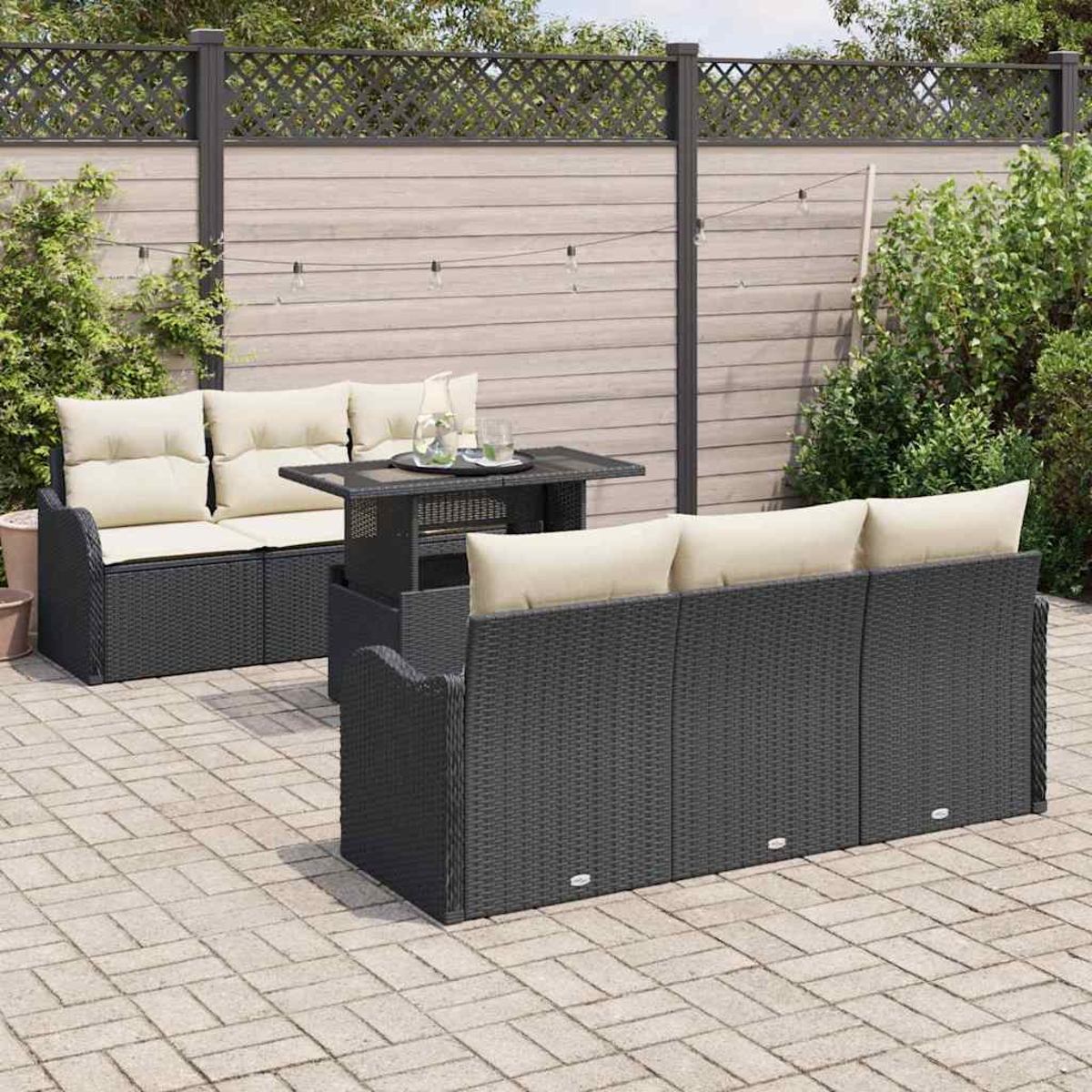 Garten-Terrasseneinrichtung mit dunklen Korbmöbeln, beigen Kissen, einem Tisch und einem Holzzaun im Hintergrund. Gepflasterter Boden.