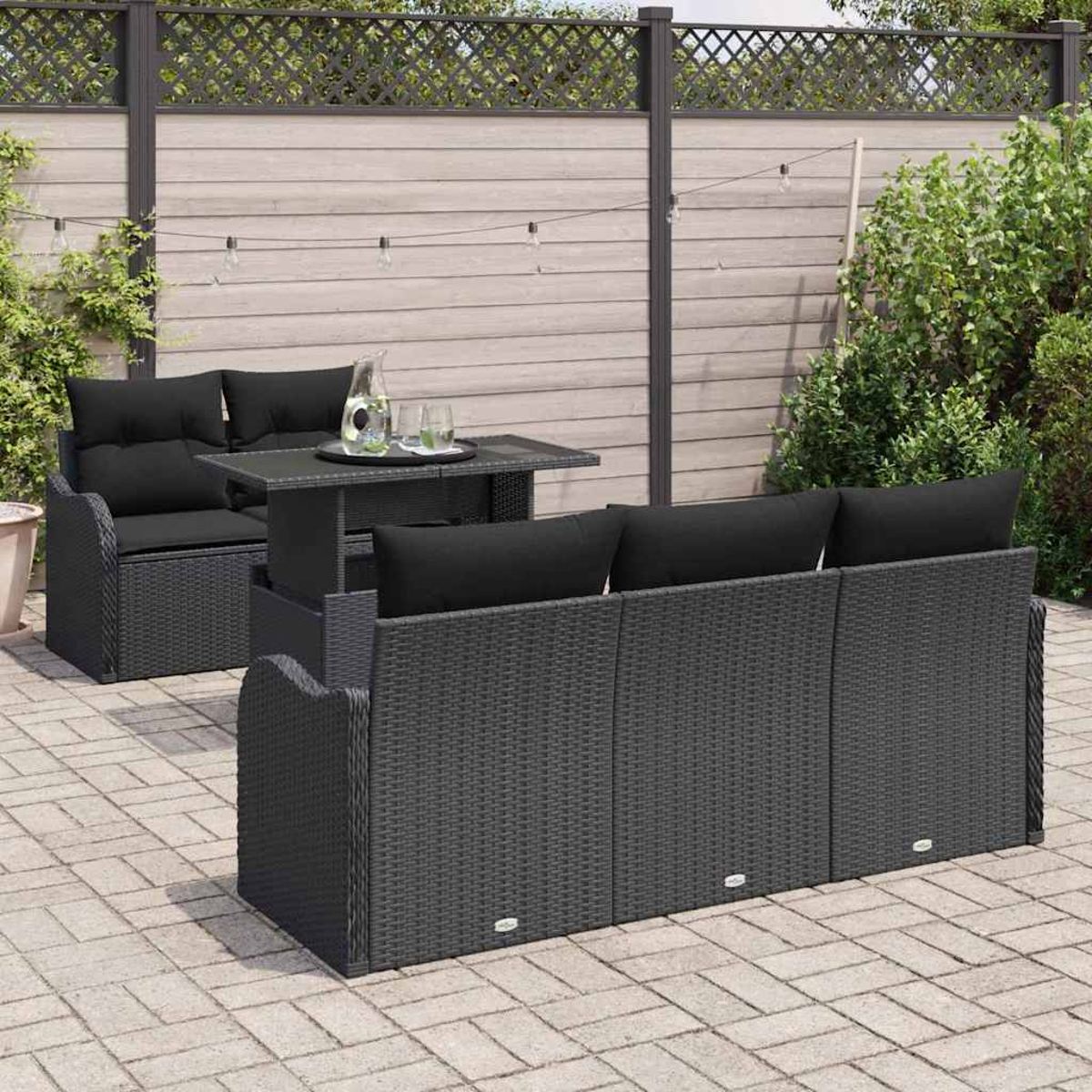 Gartenmöbel-Set: Schwarzes Korbmöbel, Tisch, schwarze Kissen, Hintergrund: Zaun und Pflanzen.