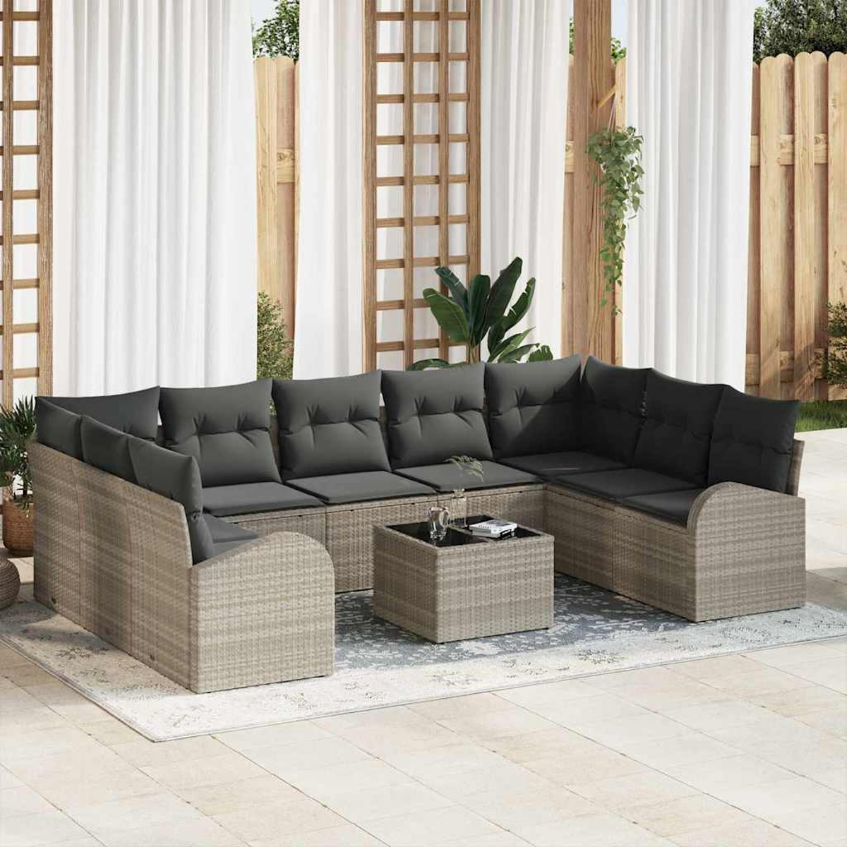 Gartensektionssofa mit grauen Kissen, Couchtisch und Vorhängen. Auf einem Teppich, mit einem Holzzaun im Hintergrund.