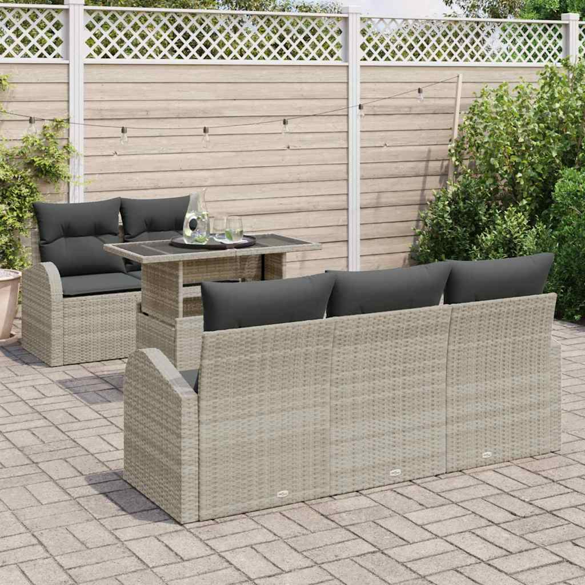 Gartenmöbel-Set mit Korbmöbeln, grauen Kissen und einem Tisch. Hintergrund mit Holzzaun und Grün, auf Steinterrasse.