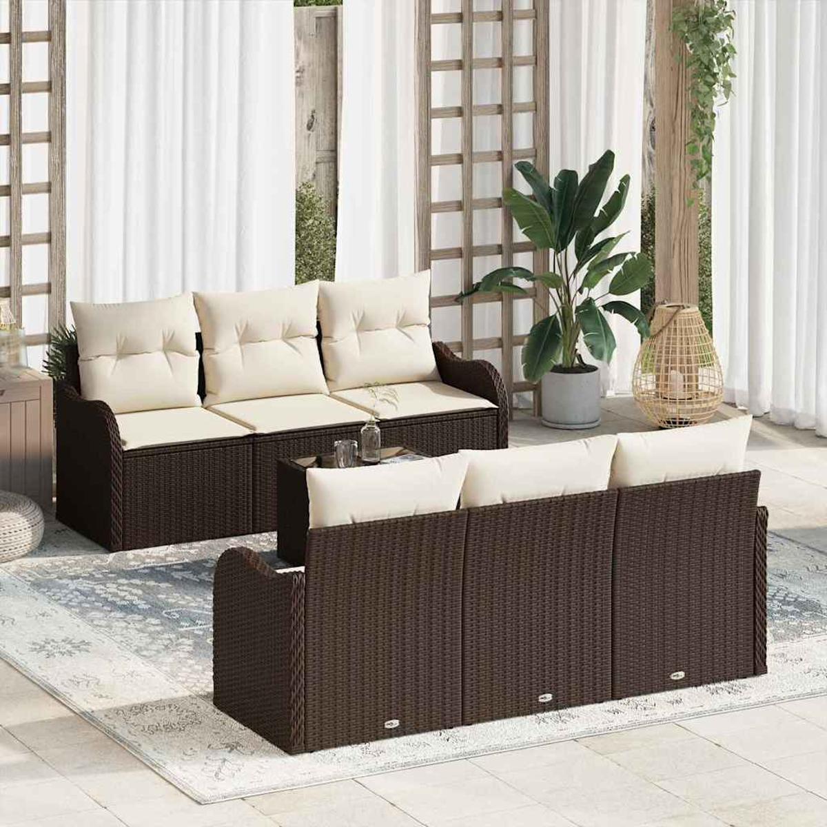 Terrassenset mit braunen Korbsesseln, cremefarbenen Kissen und einem Glastisch. Eine große Topfpflanze und eine dekorative Lampe sind ebenfalls sichtbar.
