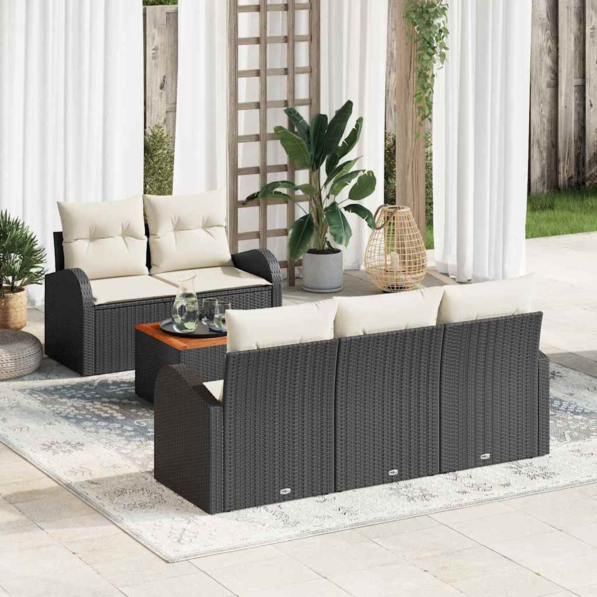 Terrassengarnitur mit schwarzem Korbmöbeln, cremefarbenen Kissen, einem Holztisch und Pflanzen, mit weißen Vorhängen im Hintergrund.