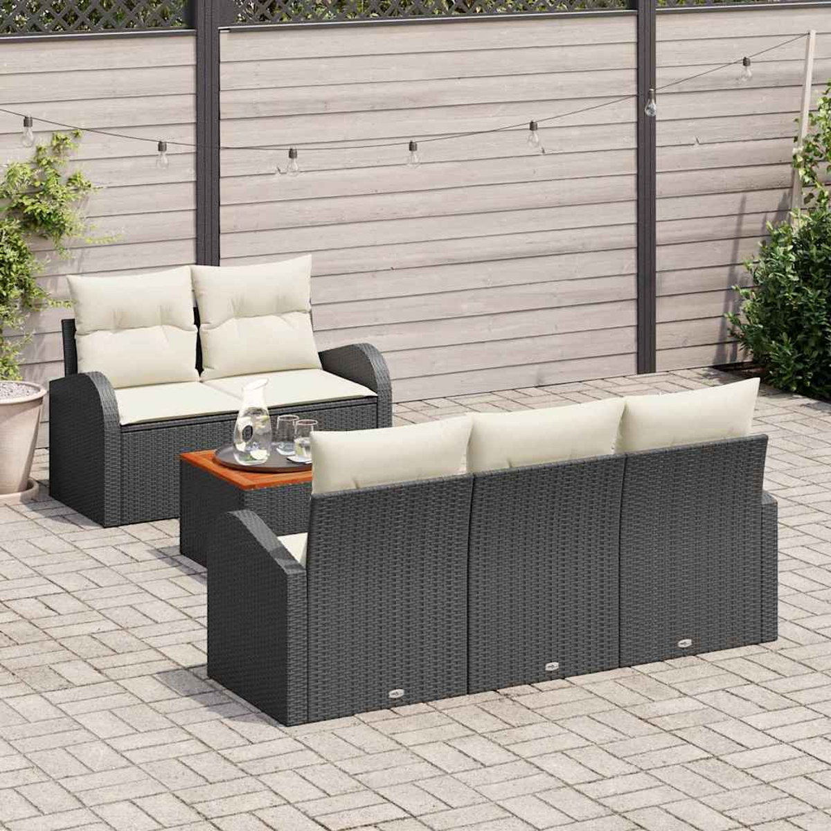 Gartenmöbel-Set: Sofa, Liebesitz und Couchtisch. Hintergrund: Holzzaun, Lichterkette und Pflanzen. Graues gewebtes Material.
