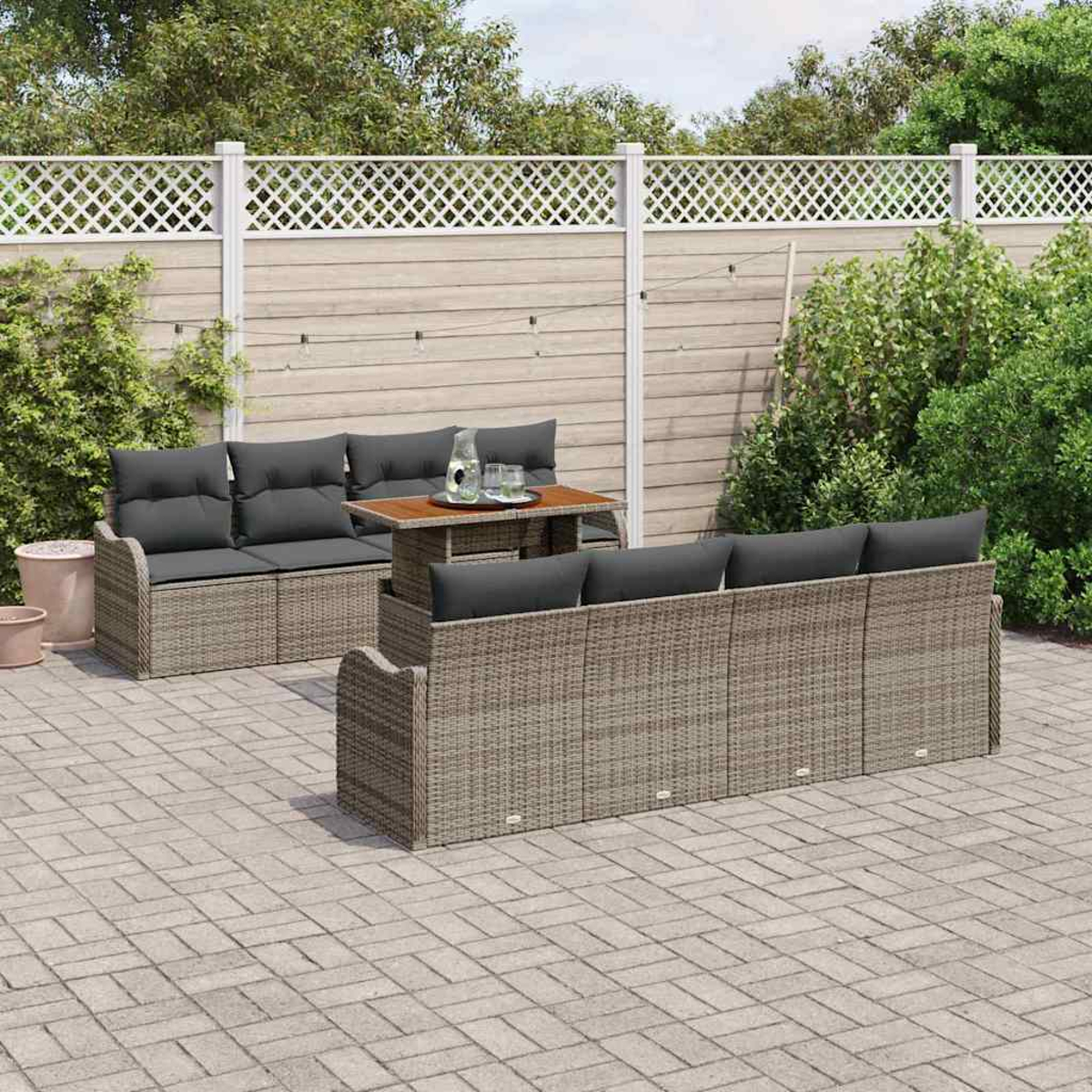 Conjunto de patio al aire libre con sofá y sillón de mimbre, cojines grises y mesa de madera. Valla blanca y plantas verdes al fondo.
