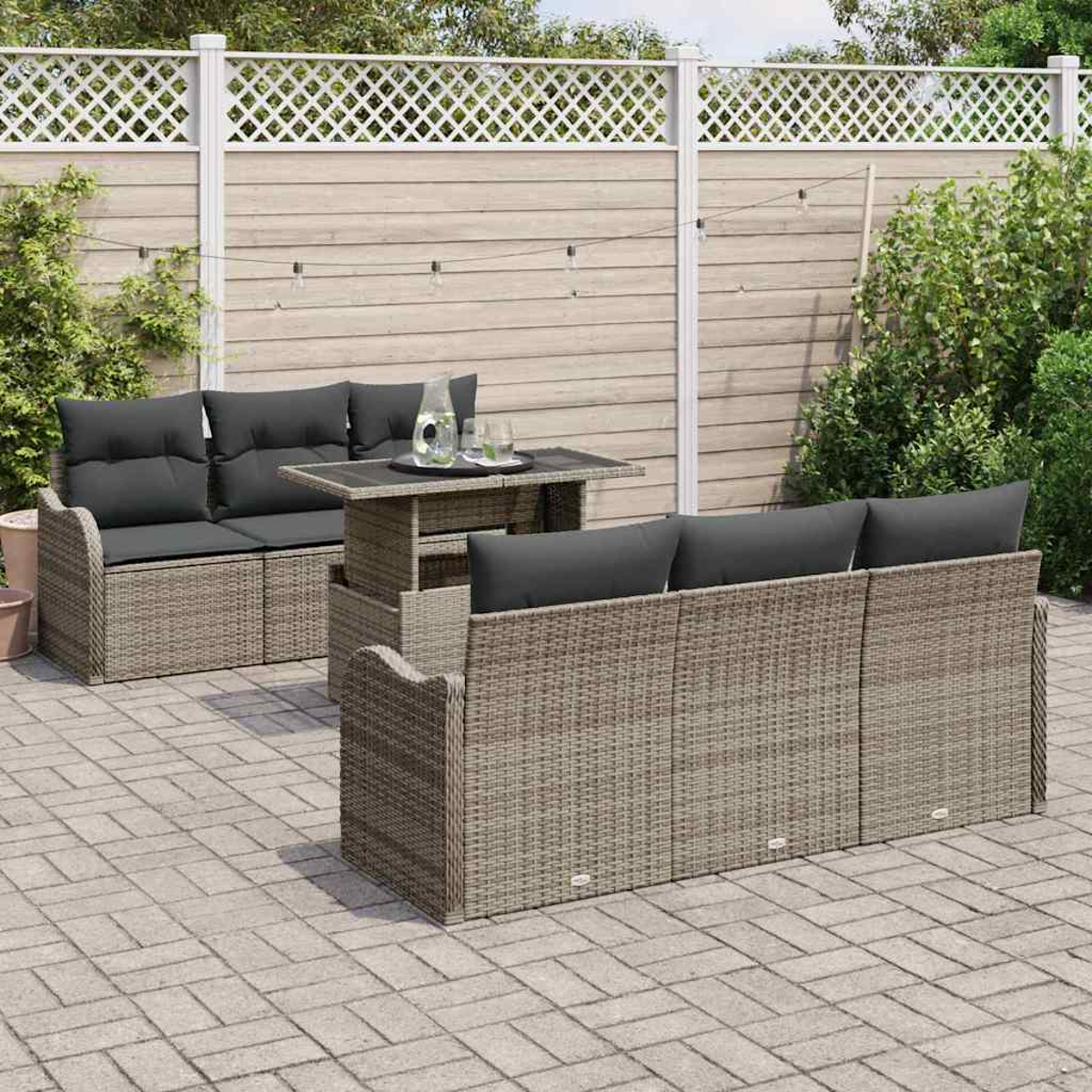 Juego de muebles de mimbre para exteriores con cojines grises, mesa con bebidas, en patio de piedra. Valla blanca y vegetación al fondo.